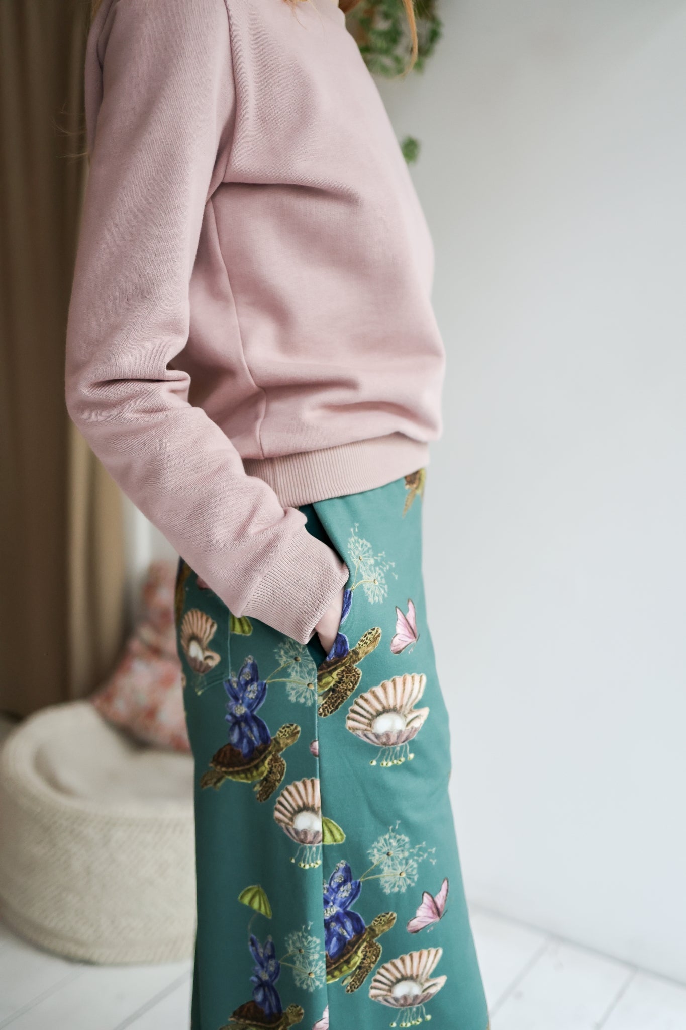 Woman wearing a pink sweater and teal floral pants in a room with a blurred background.
