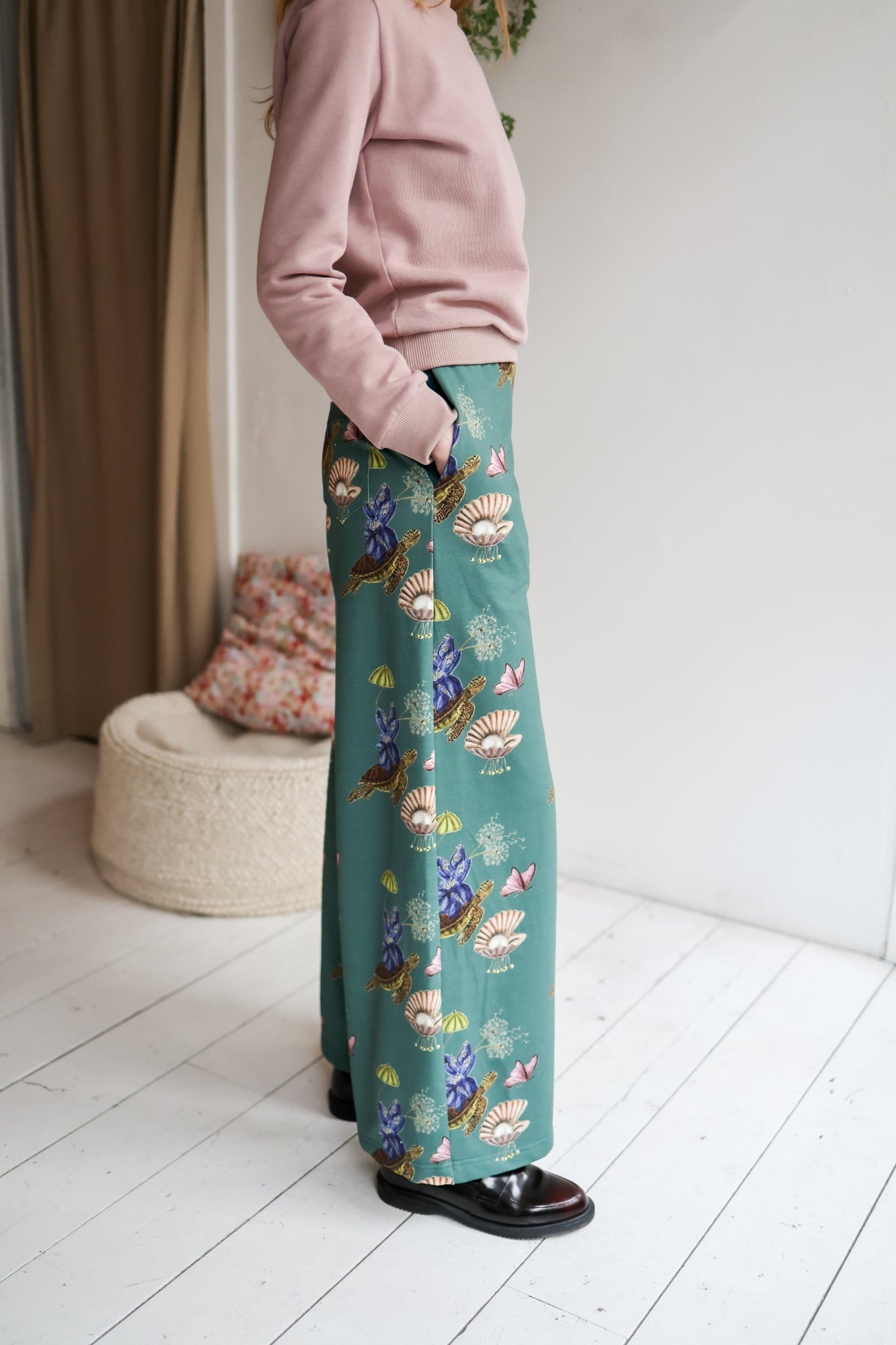 Woman wearing a pink sweater and teal floral pants in a room with a white wall and wooden floor.