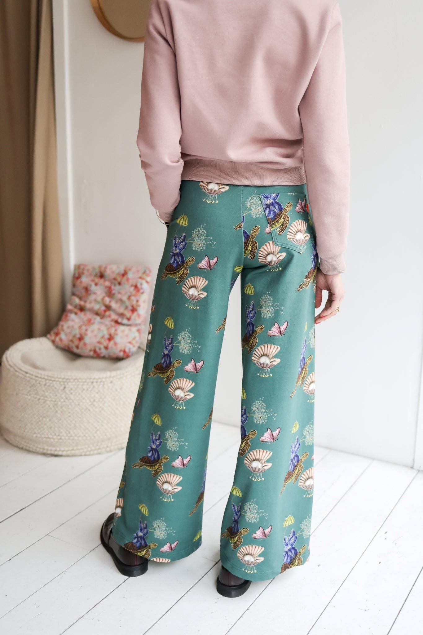 Woman wearing a pink sweater and teal floral pants in a room with a white wall and wooden floor.