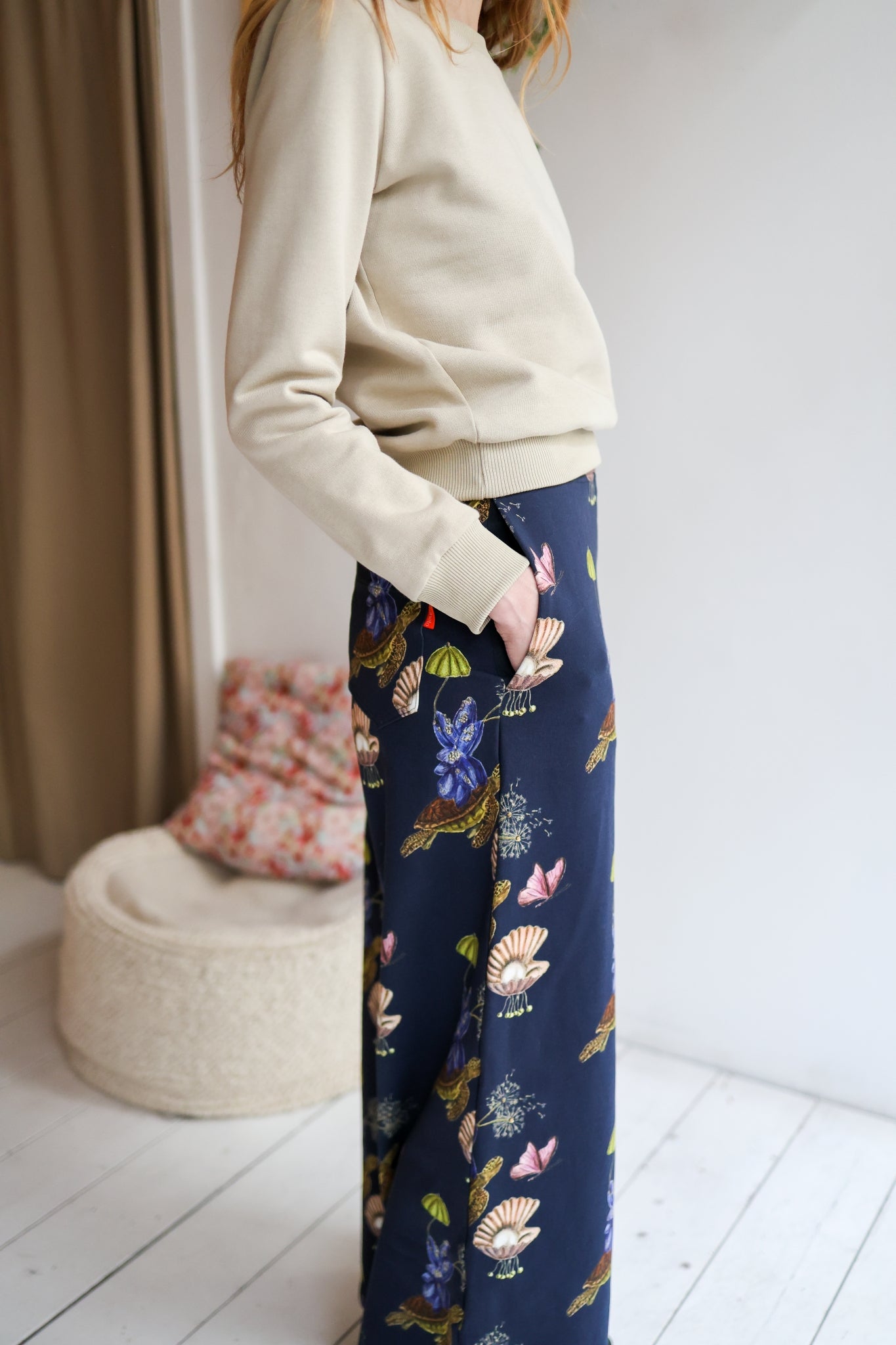 Woman wearing a sweater and patterned pants in a room with a white wall and wooden floor.