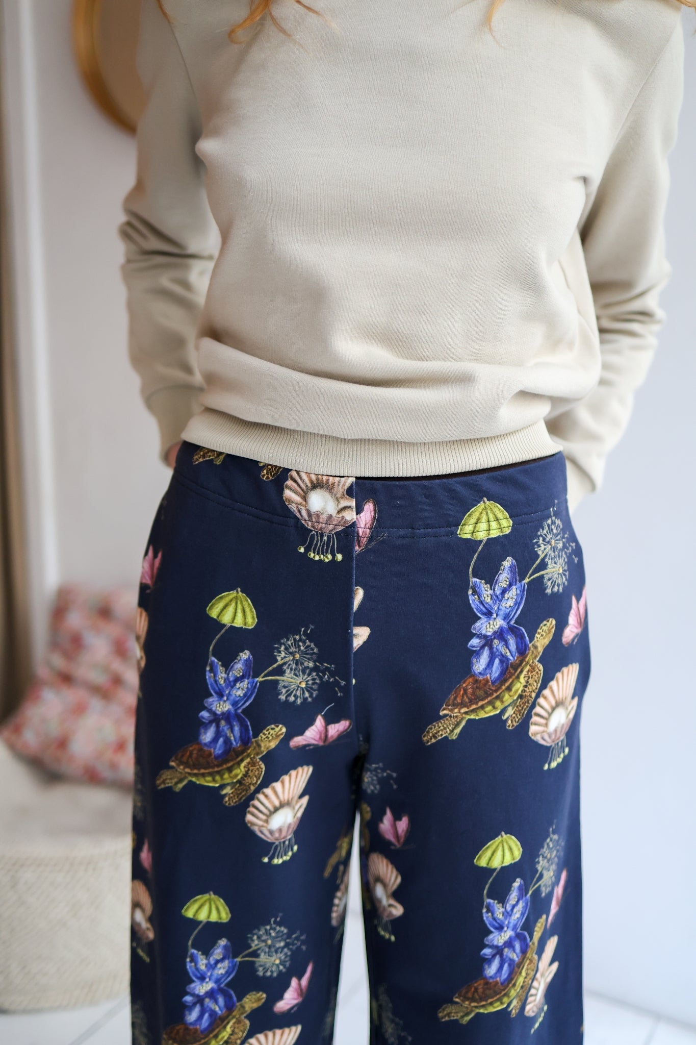 Woman wearing a sweater and patterned pants in a room with a blurred background.