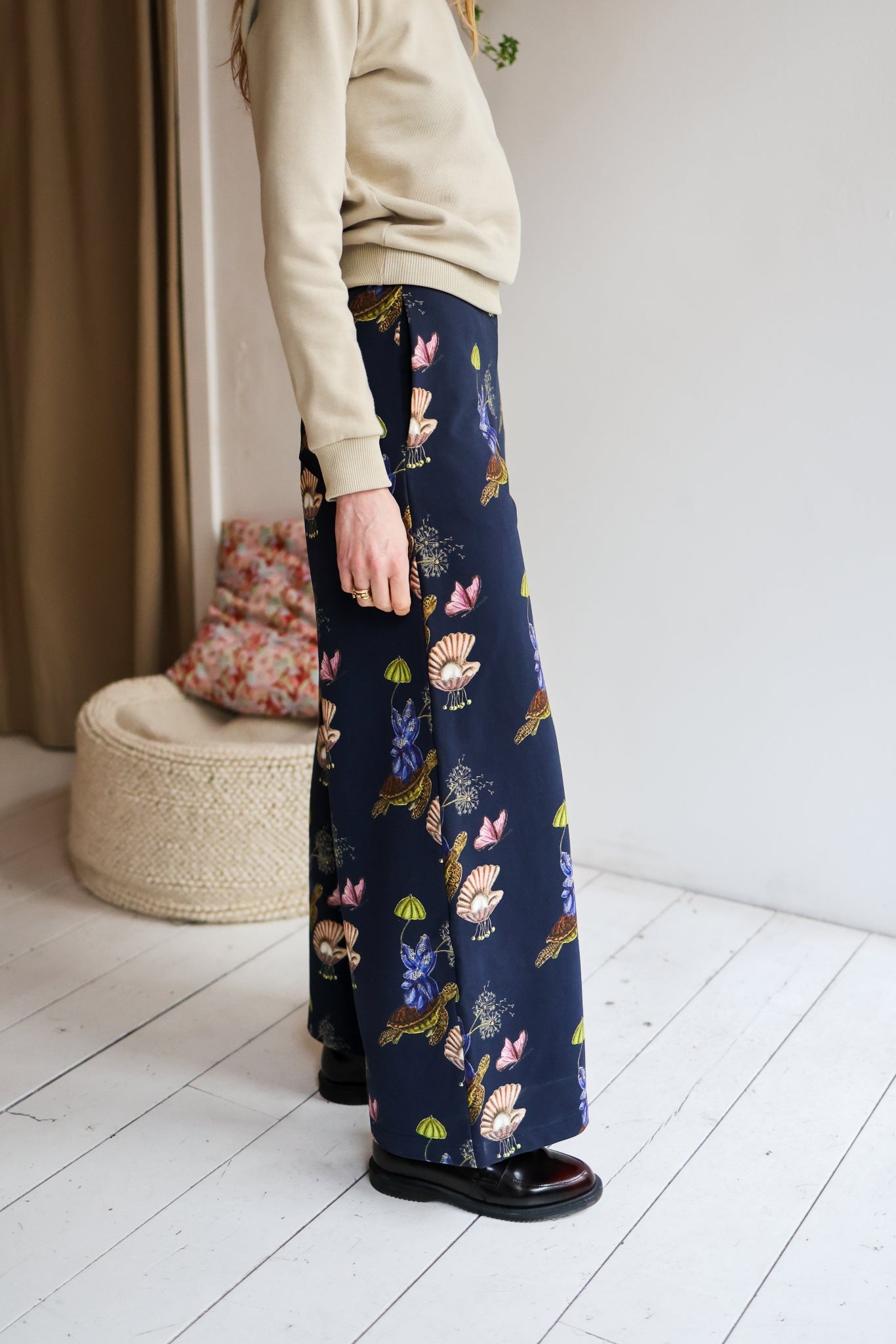 Woman wearing a sweater and patterned pants in a room with a white wall and wooden floor.
