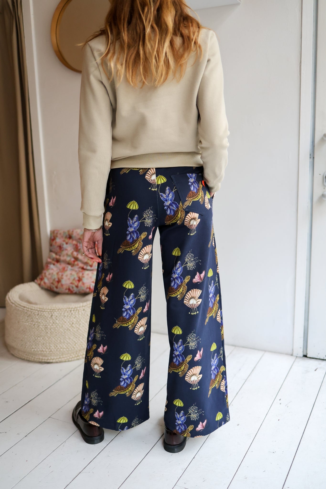 Woman wearing a sweater and patterned pants in a room with a white wall and wooden floor.