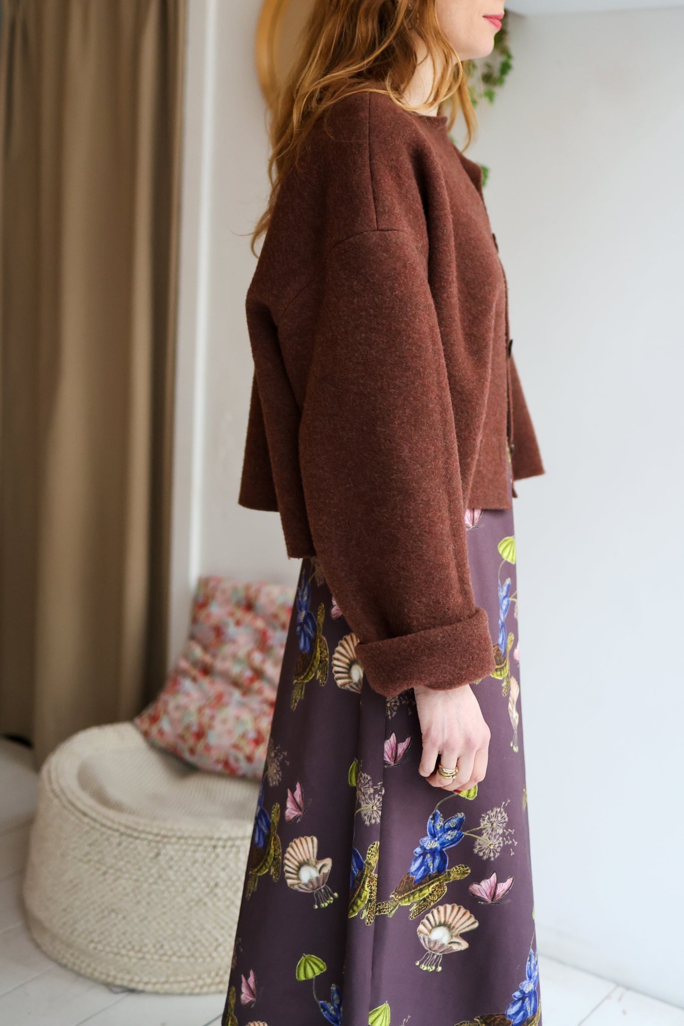 Woman wearing a brown jacket over a patterned dress in a room with a blurred background.