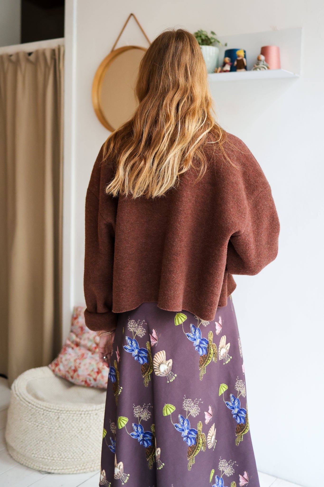 Woman wearing a brown jacket over a patterned dress in a room with a white wall.