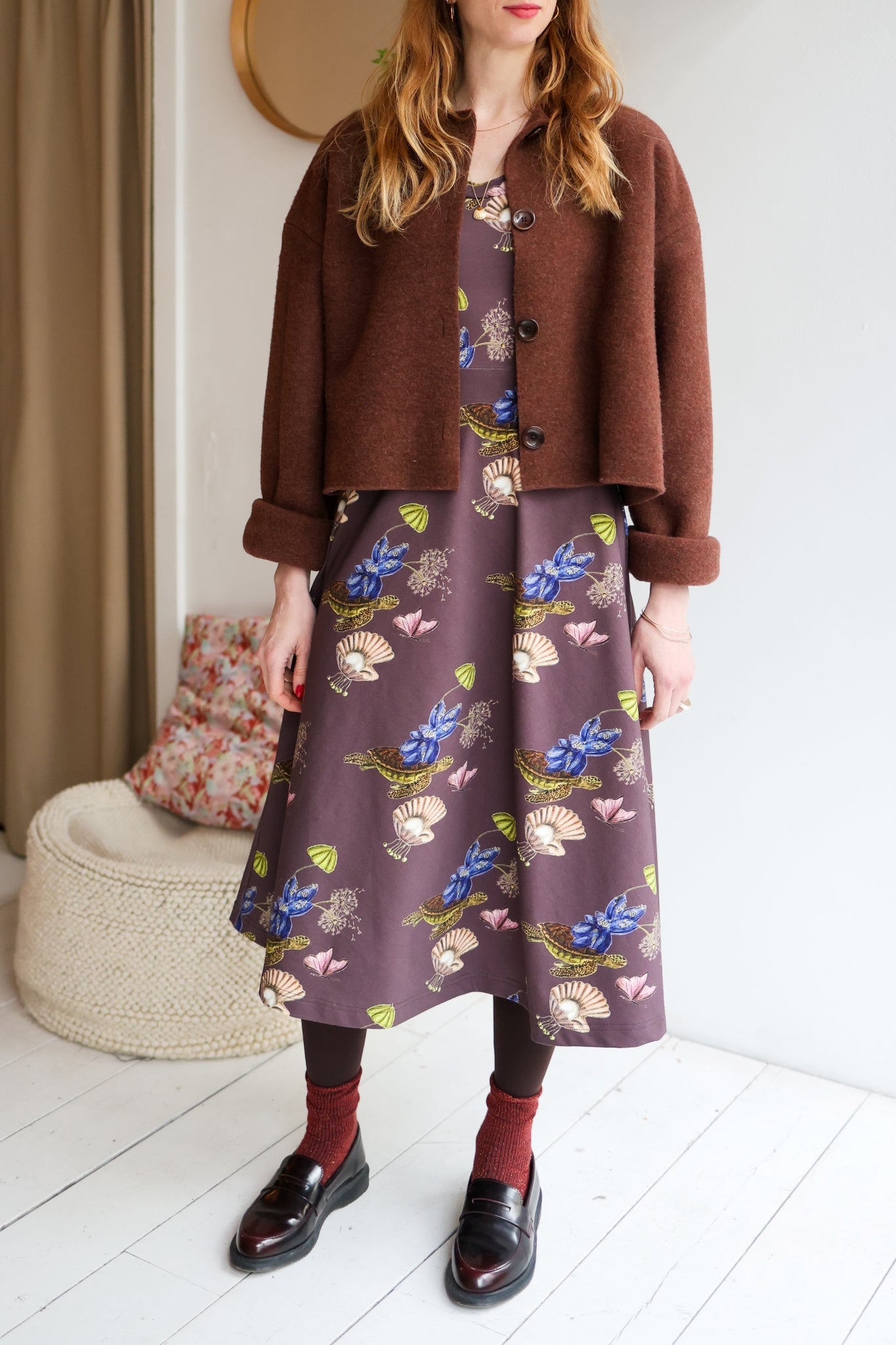 Woman wearing a brown jacket and patterned dress with dark shoes indoors.