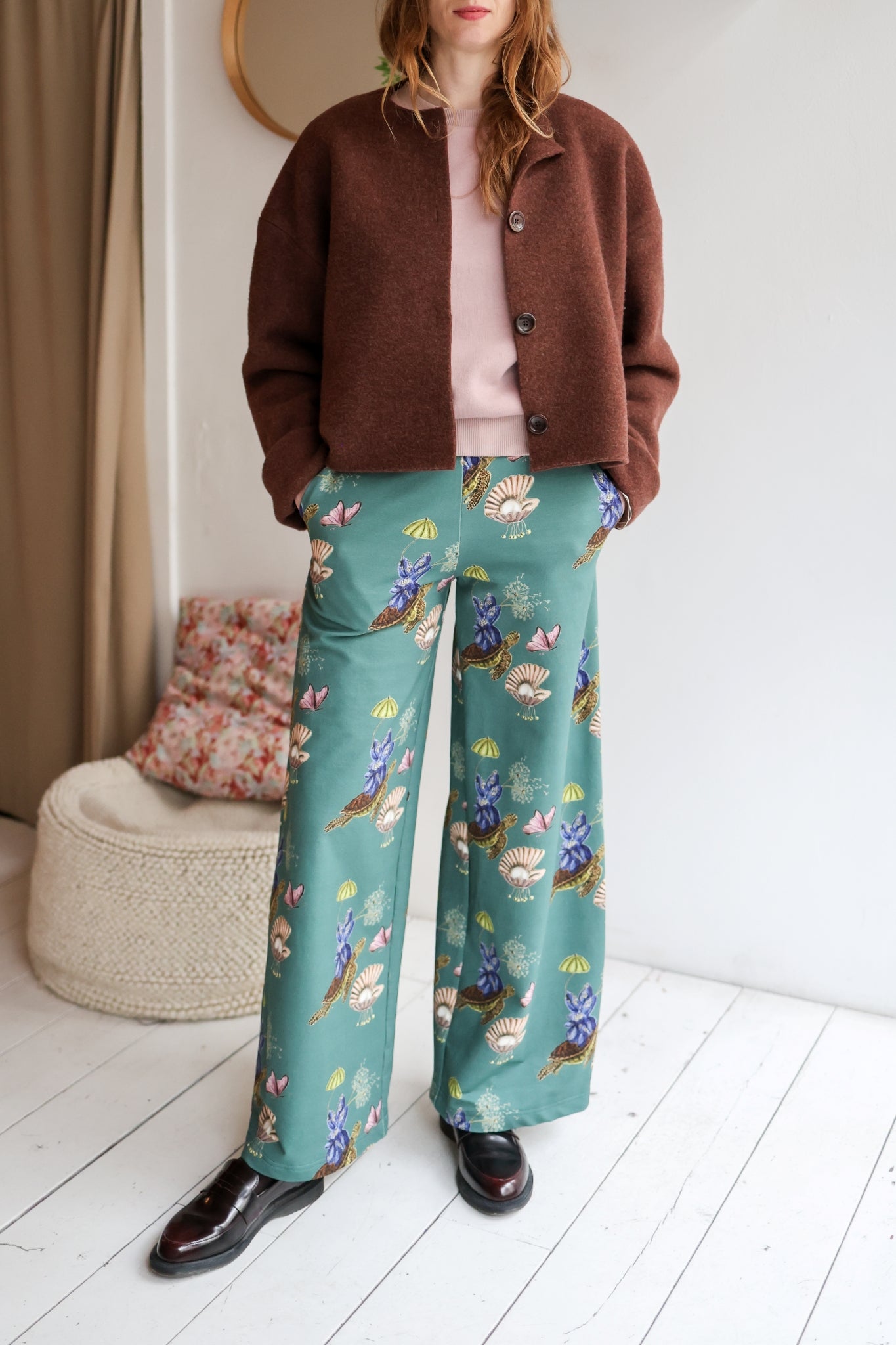 woman wearing a brown jacket and patterned teal pants indoors.