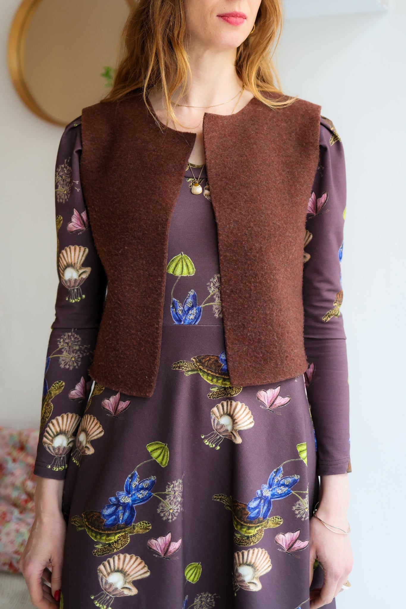 Woman wearing a brown vest over a patterned dress with a blurred background.