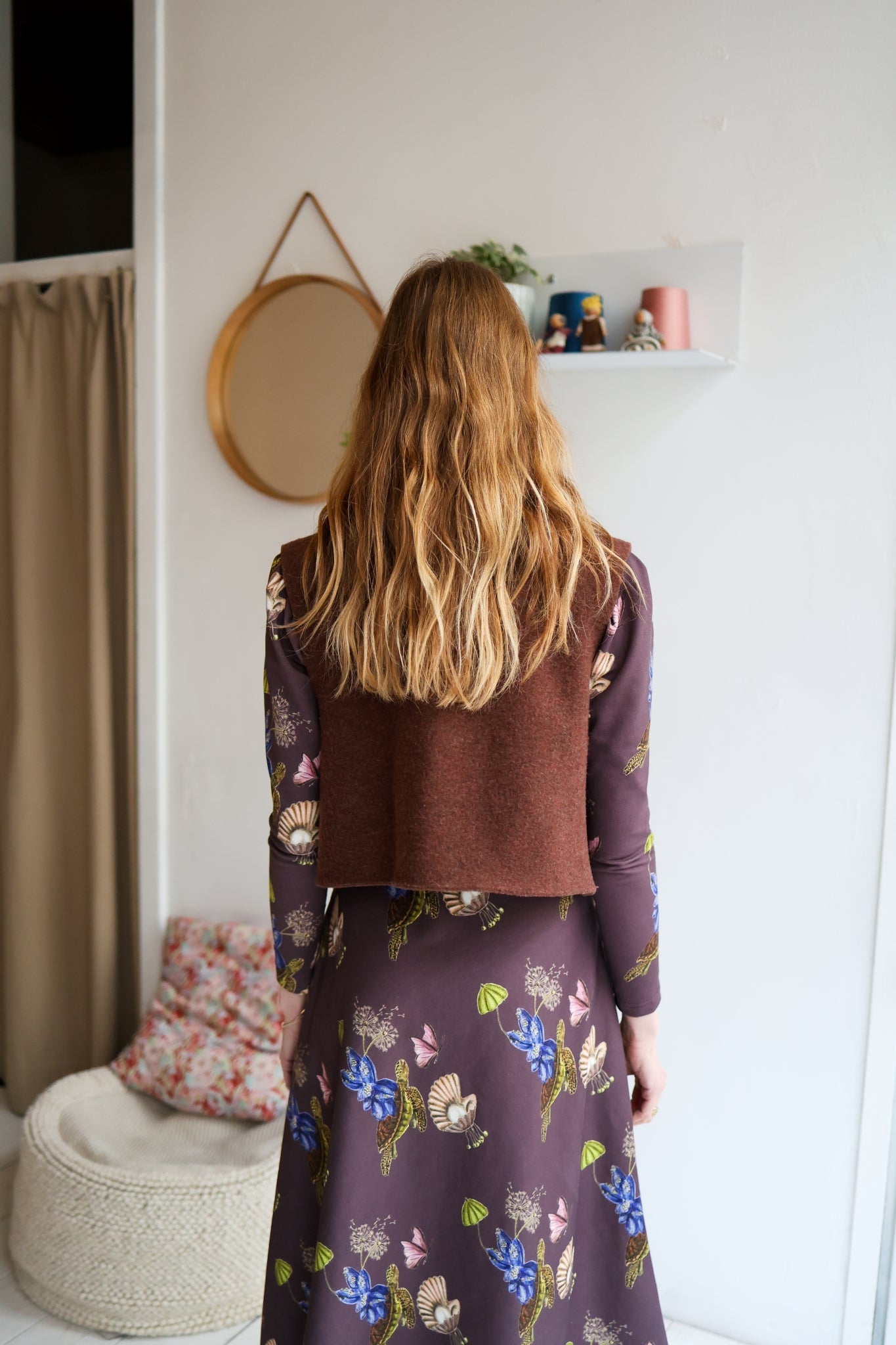 Woman wearing a brown vest over a patterned dress in a room with a white wall.