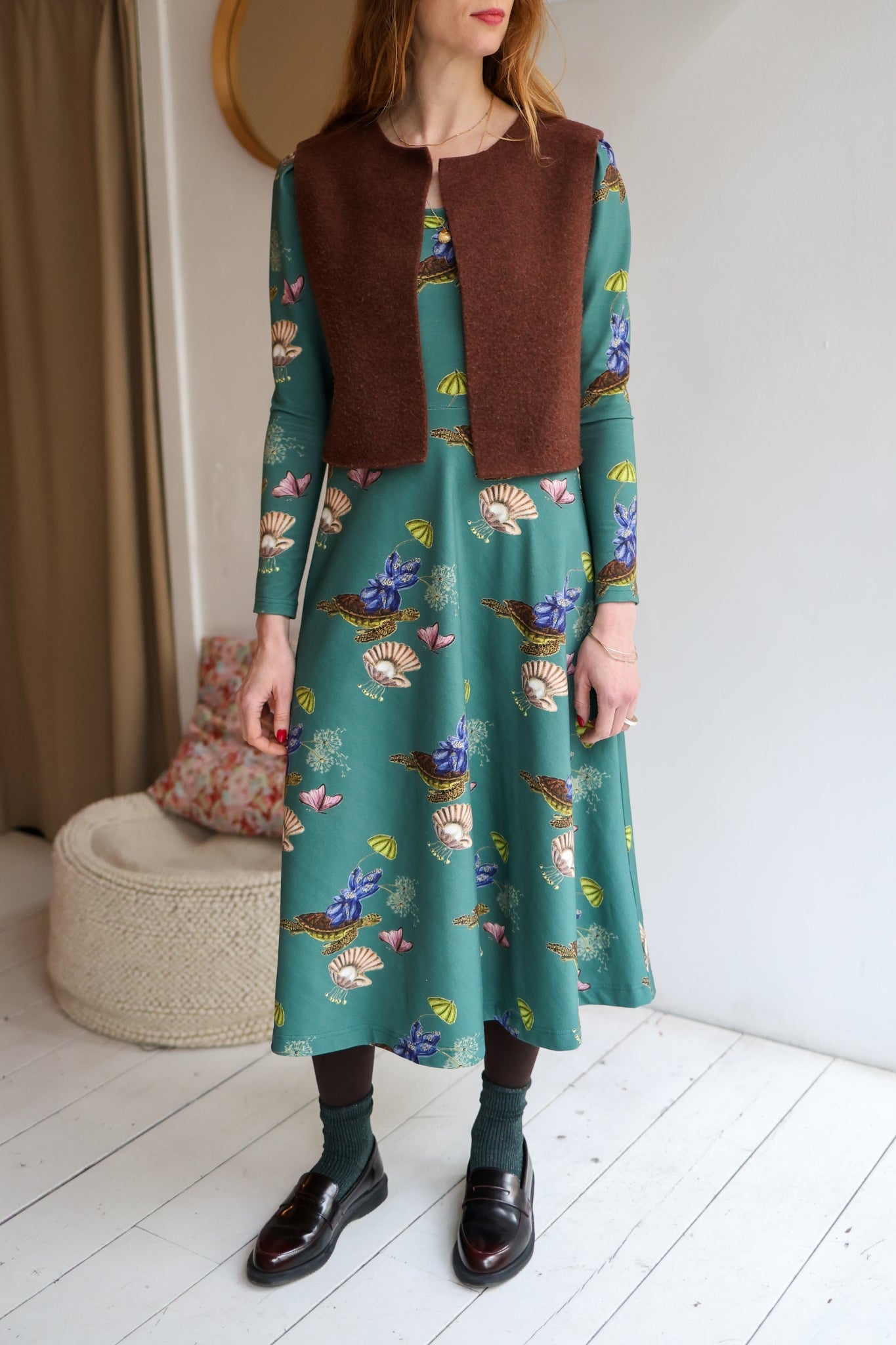 Woman wearing a brown vest over a patterned dress in a room with a white wall.