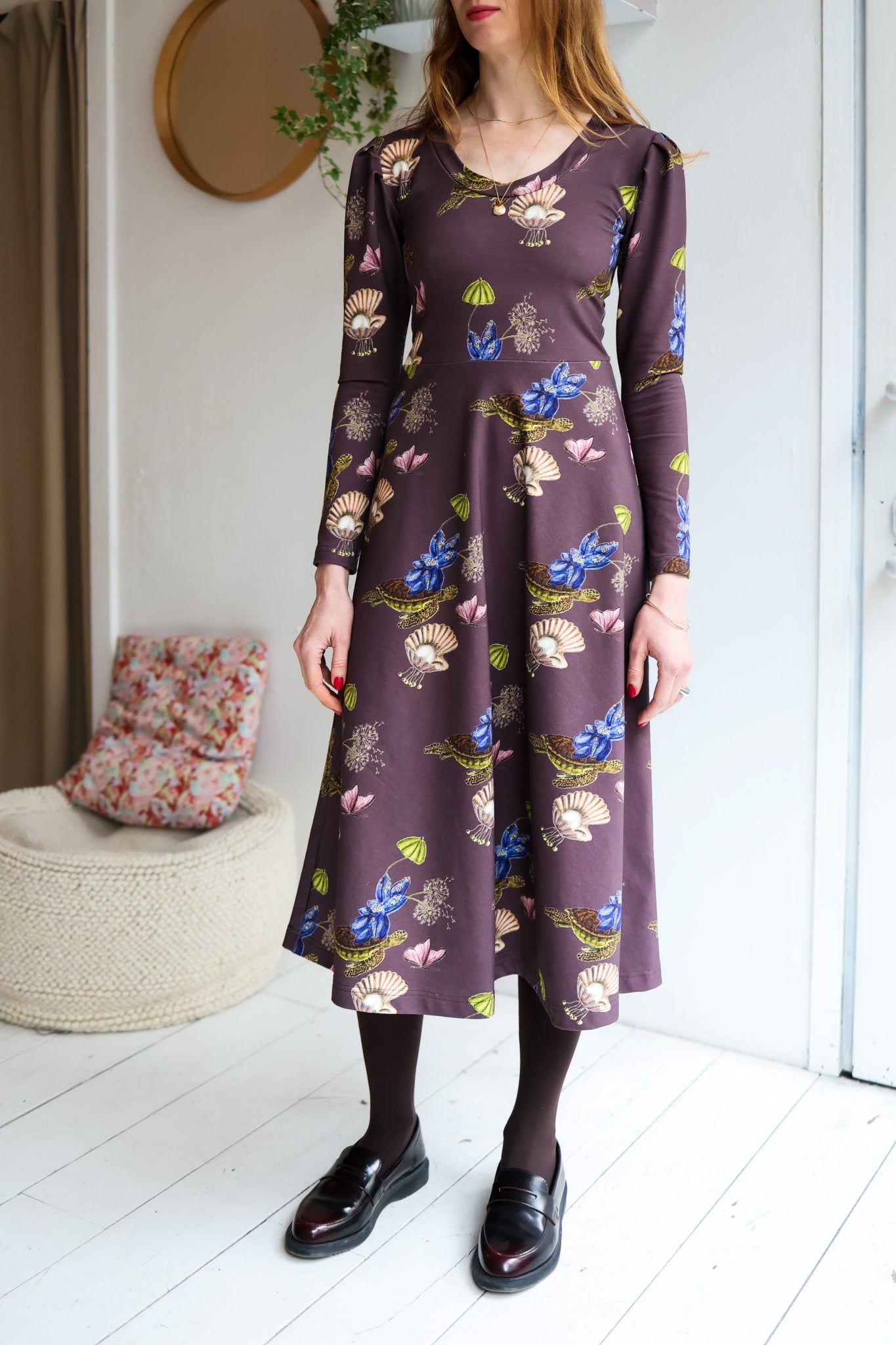Woman wearing a patterned dress with long sleeves in a room with a white wall.