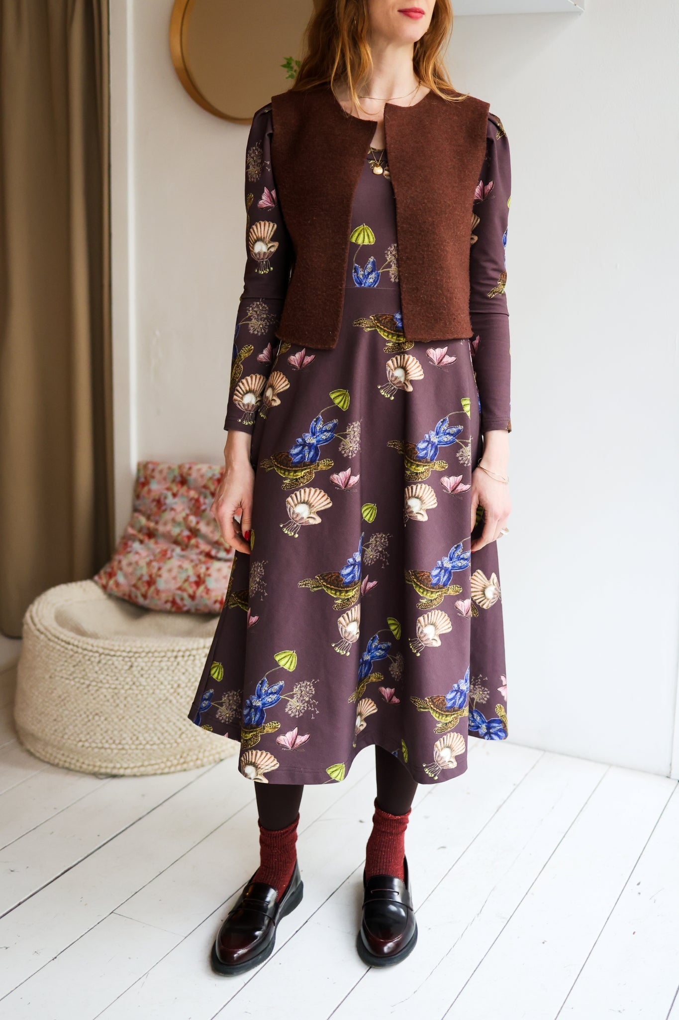 Woman wearing a patterned dress and brown vest in a room with a white floor and beige curtain.