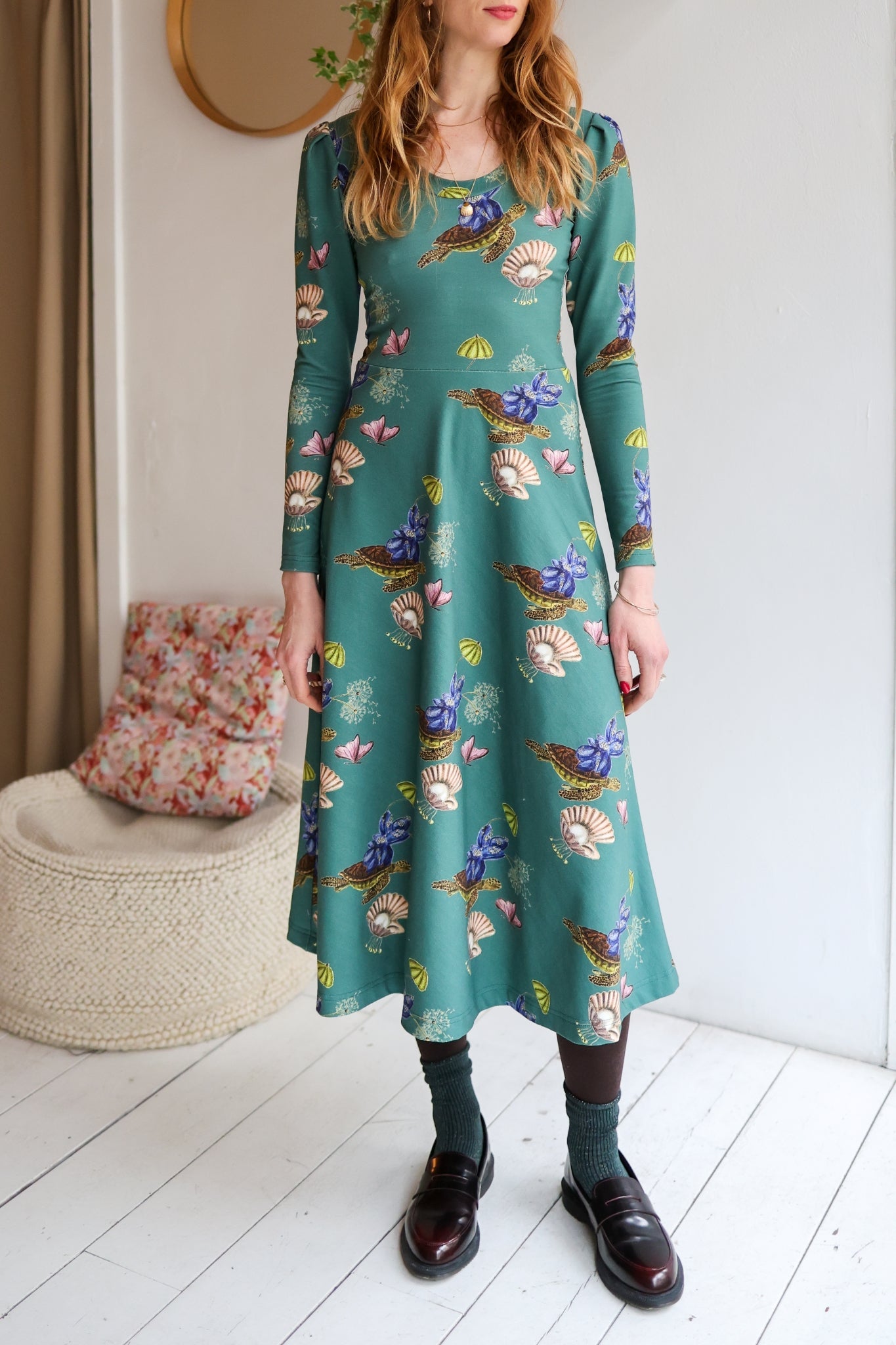 Woman wearing a patterned dress standing in a room with a white wall and wooden floor.