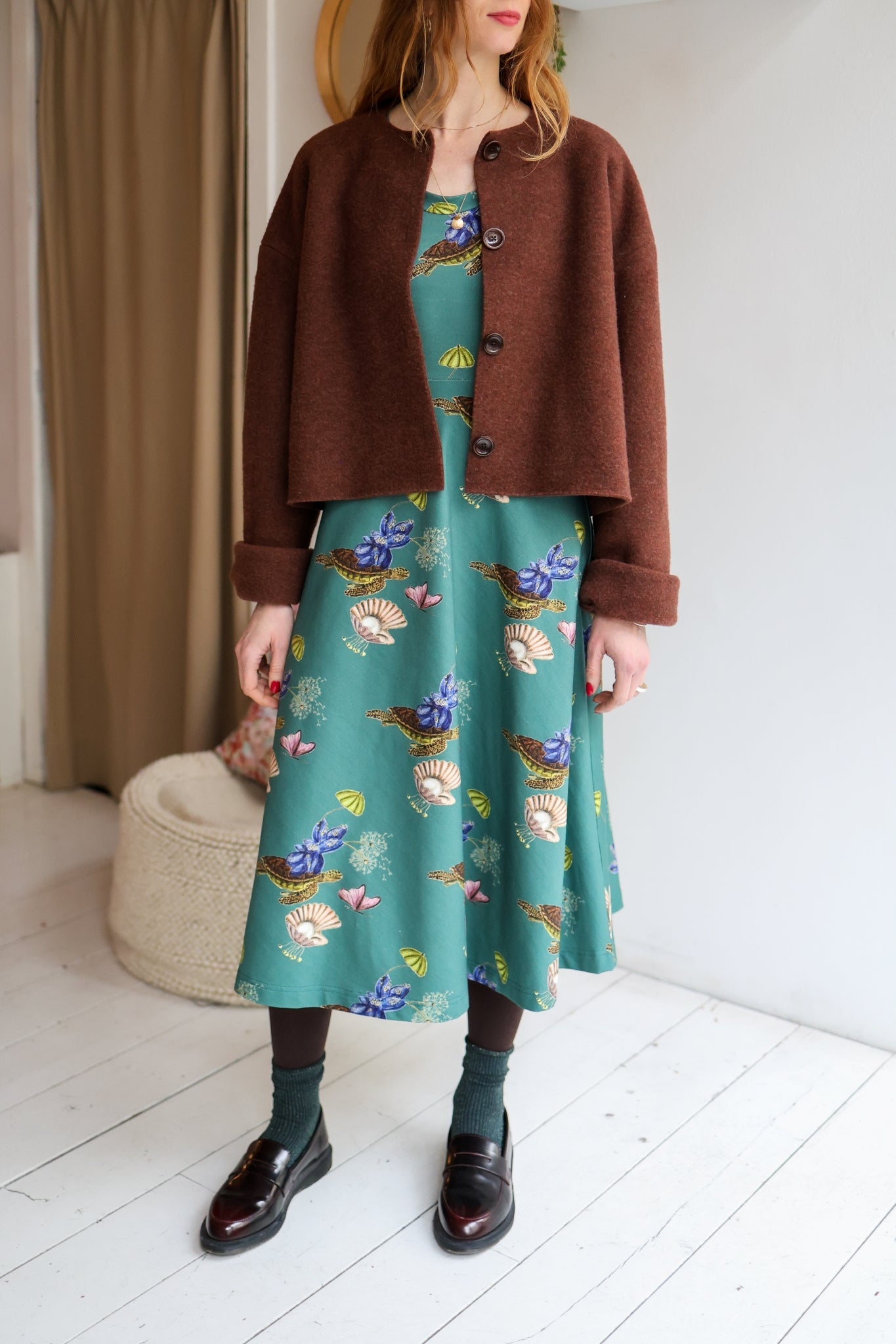 Woman wearing a brown coat over a patterned dress in a minimal indoor setting.