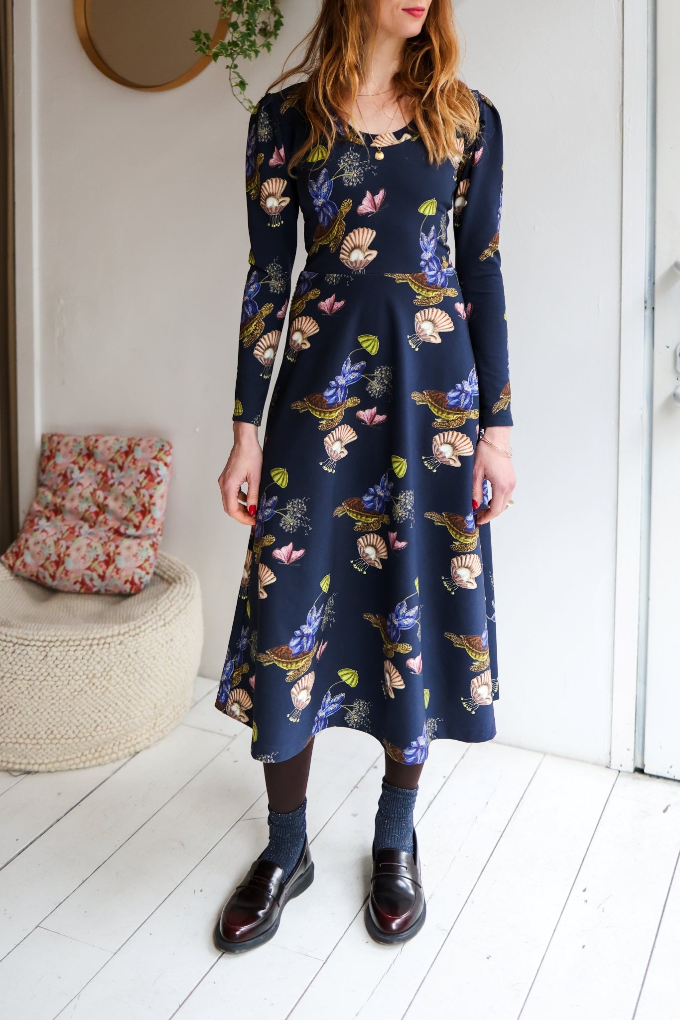 Woman wearing a floral dress standing in a room with white walls and wooden floor.