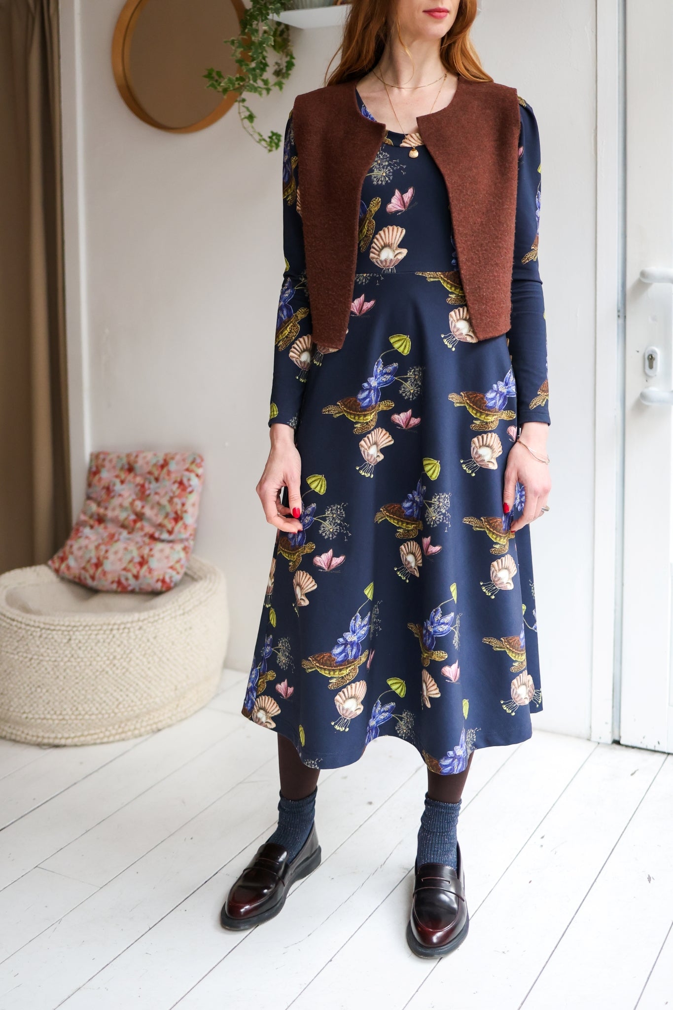 Woman wearing a floral dress with a brown vest in a room with a white floor and a plant.