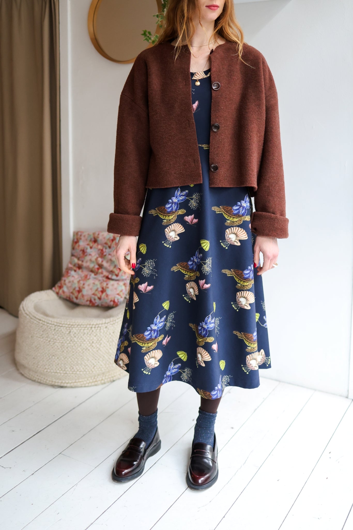 Woman wearing a brown jacket, blue floral dress, and black shoes in a room with a white wall and wooden floor.
