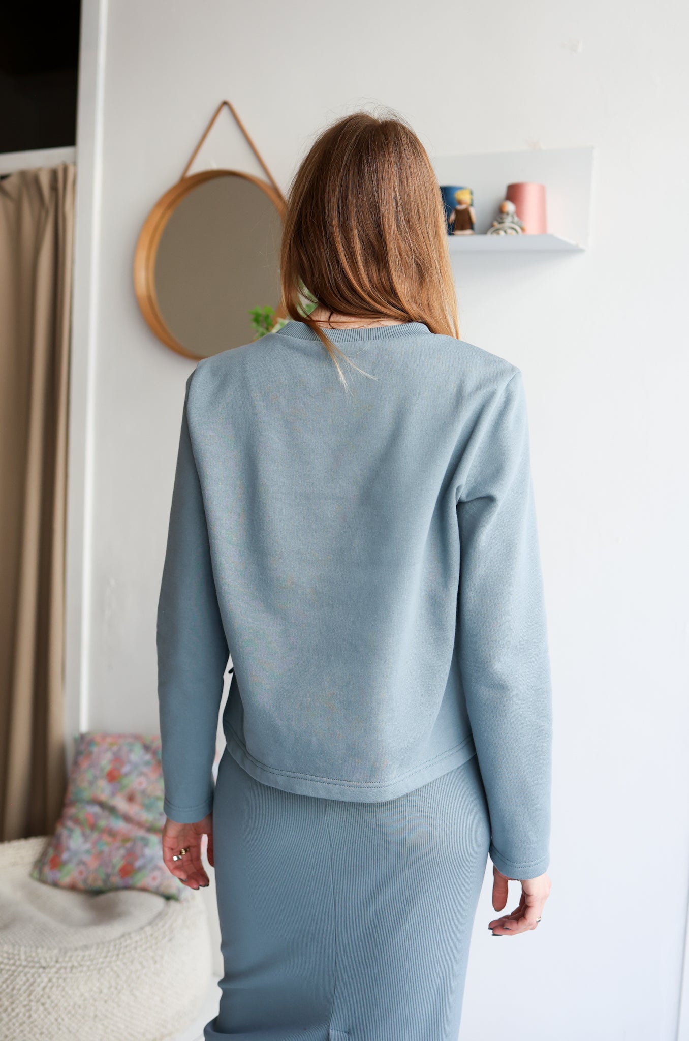 Woman wearing a light blue outfit standing in a room with a mirror and shelf.
