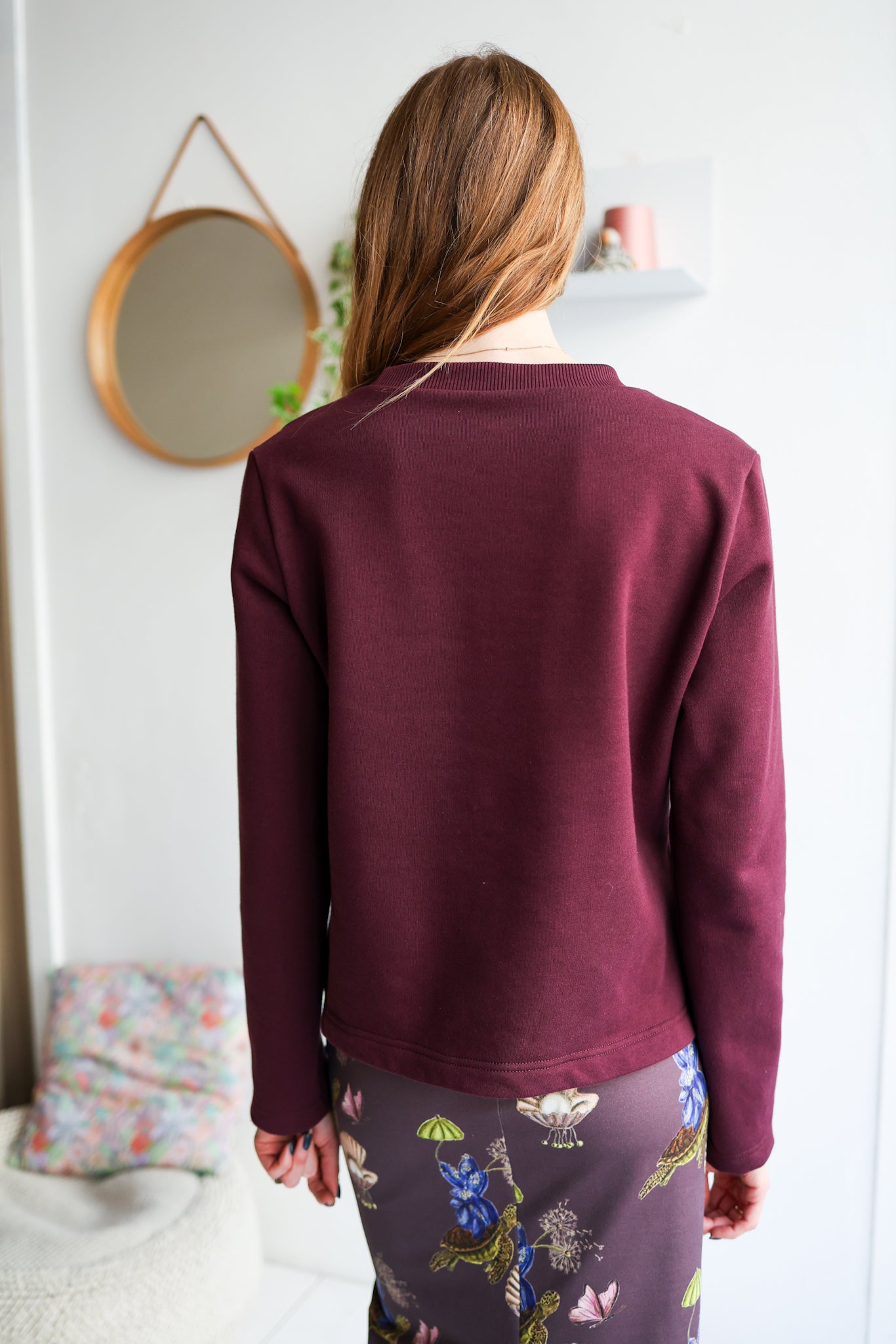 Woman wearing a bordeaux sweatshirt with a white wall and mirror in the background.