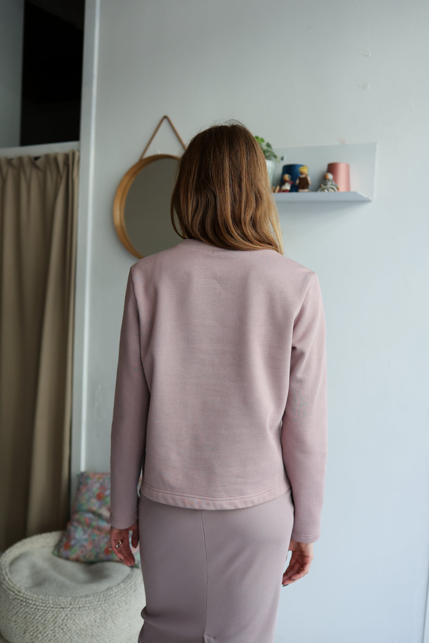 Person wearing a pink outfit standing in a room with a mirror and shelf.