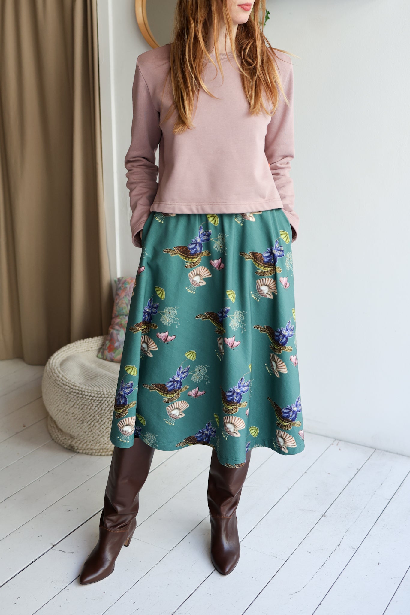Woman wearing a pink top and green floral skirt with brown boots in a room.