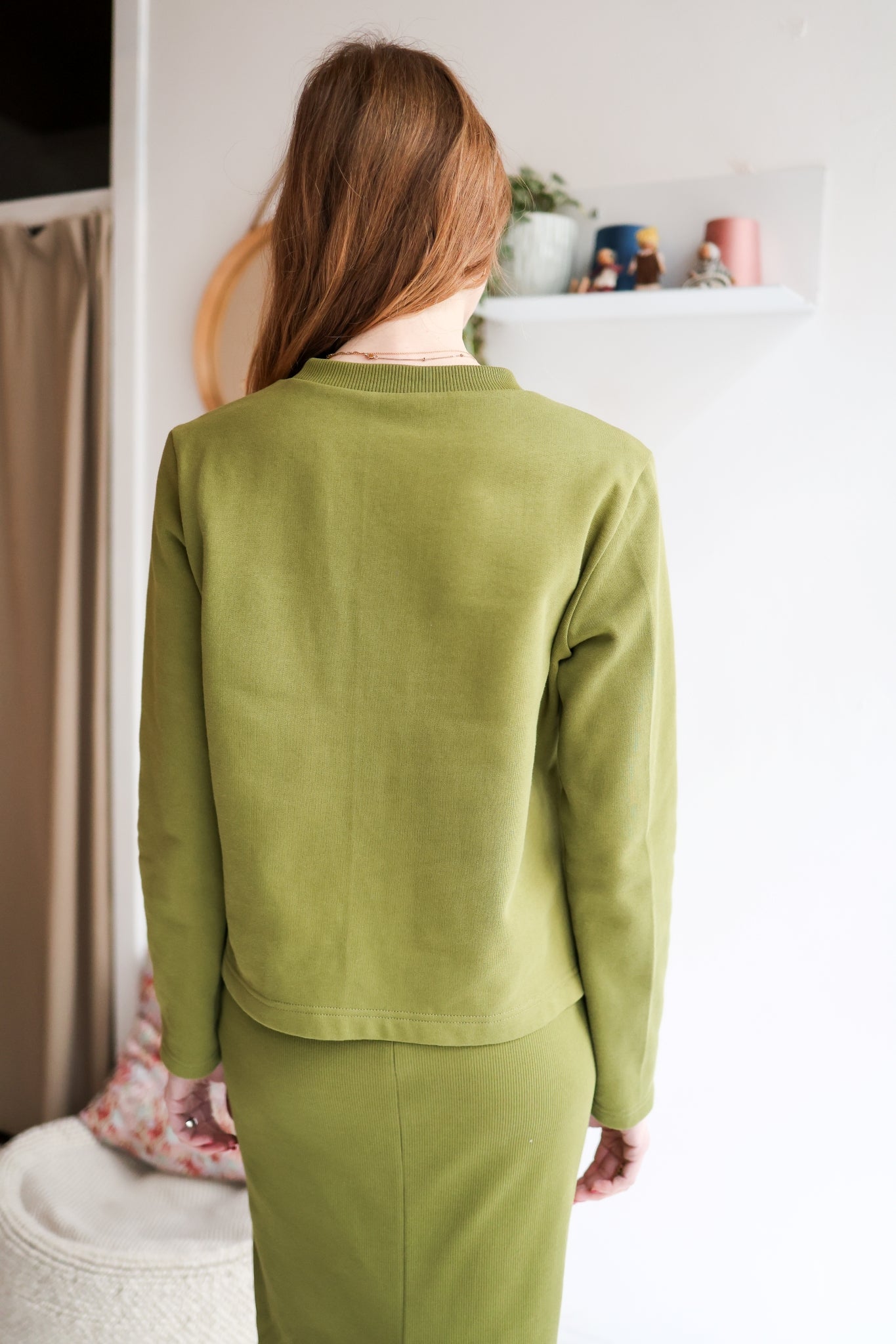 Person wearing a green outfit standing in a room with a white wall and some decor items.