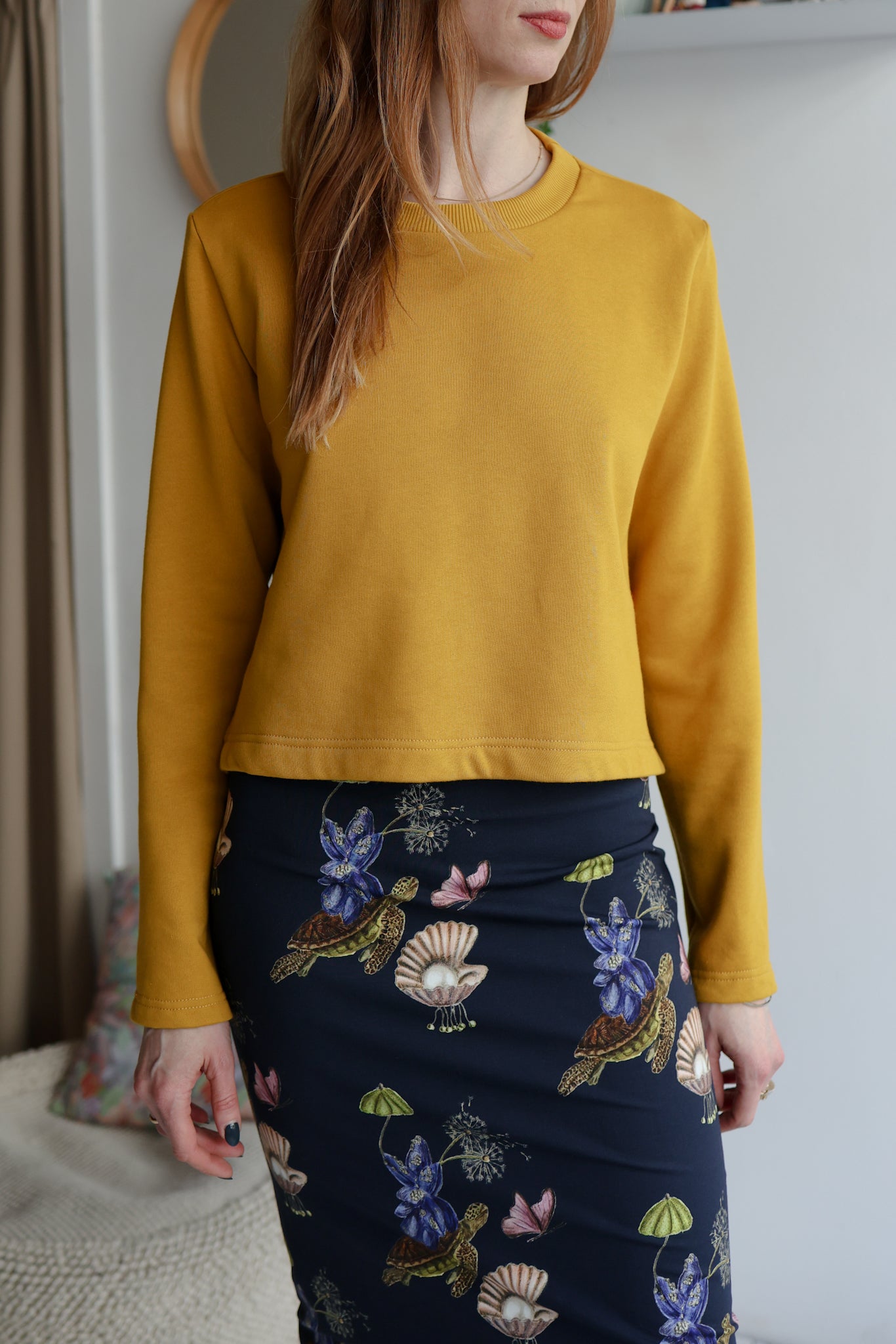 Woman wearing a yellow sweatshirt and a navy patterned skirt indoors with a blurred background.