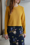 Woman wearing a yellow sweatshirt and a navy patterned skirt indoors with a blurred background.