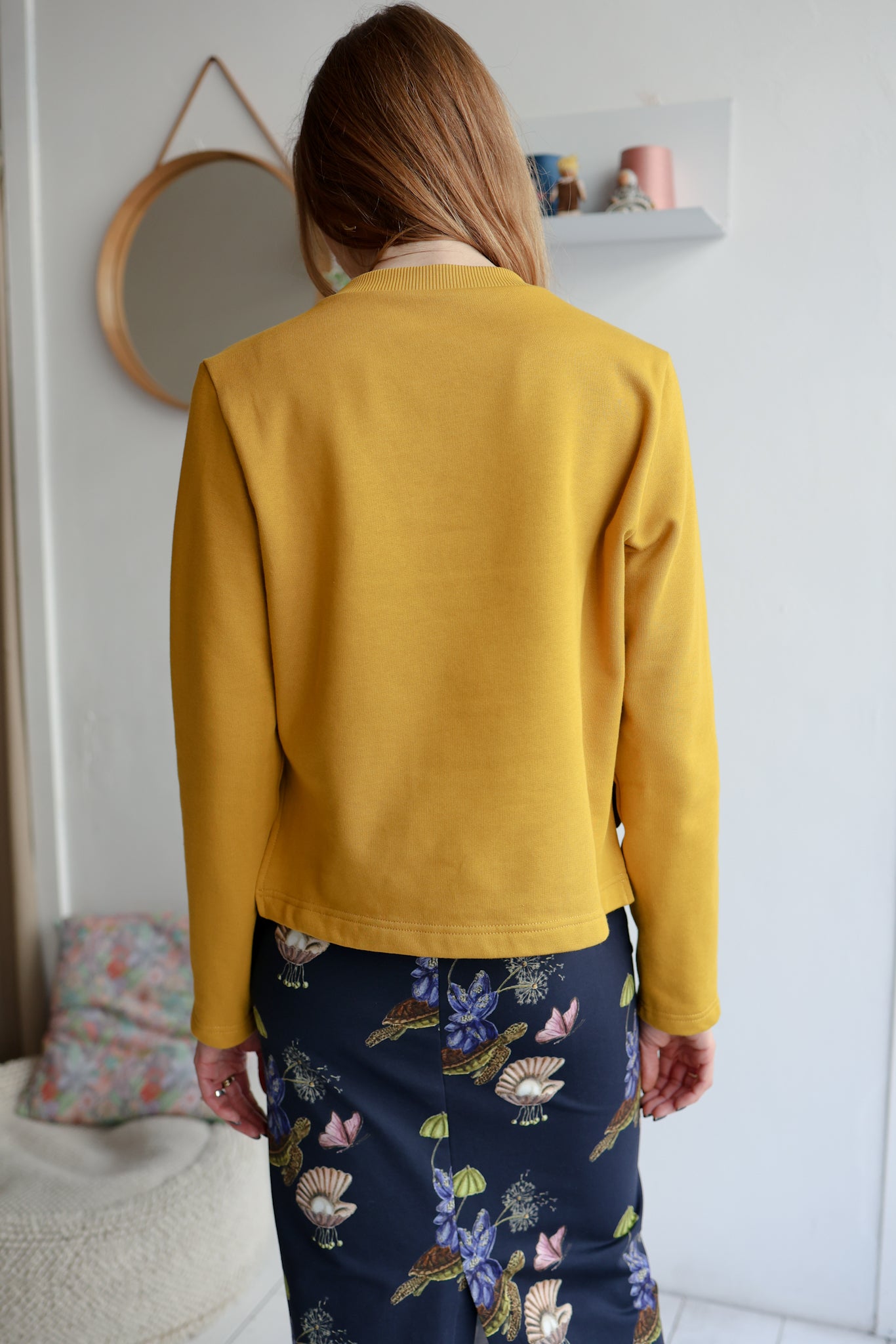 Woman  wearing a yellow sweatshirt and a navy patterned skirt standing in a room.