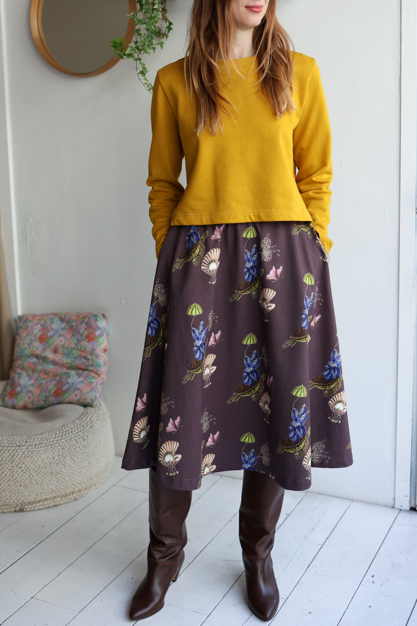 Woman wearing a yellow sweatshirt and  a navy patterned skirt n a room with white walls and a wooden floor.