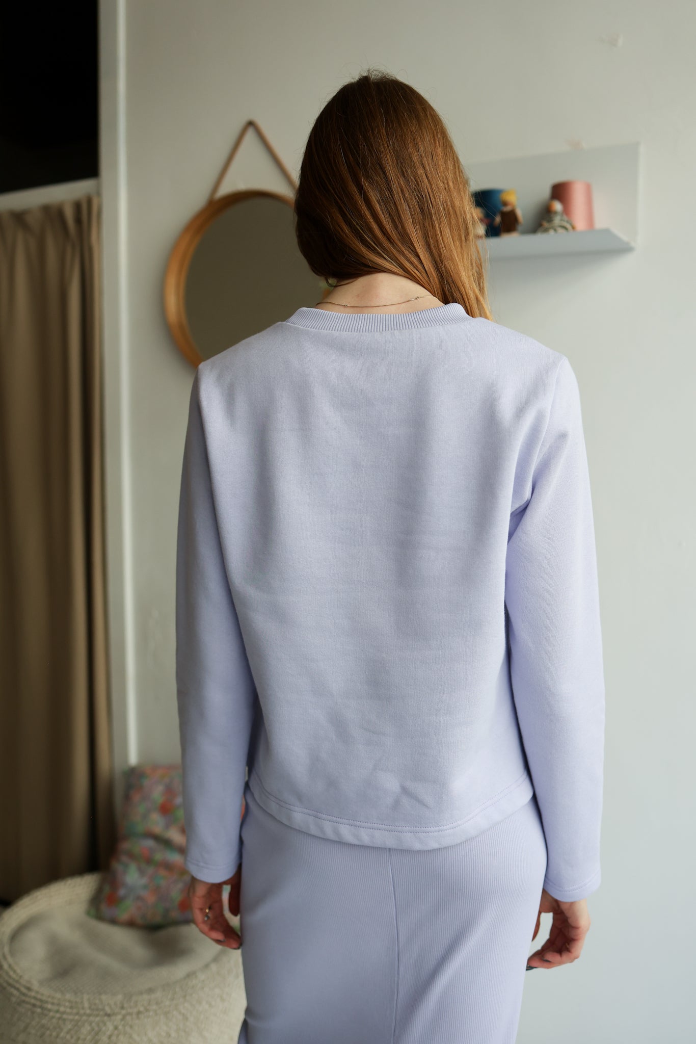 Person wearing a light purple sweatshirt and pants in a room with a mirror and shelf.