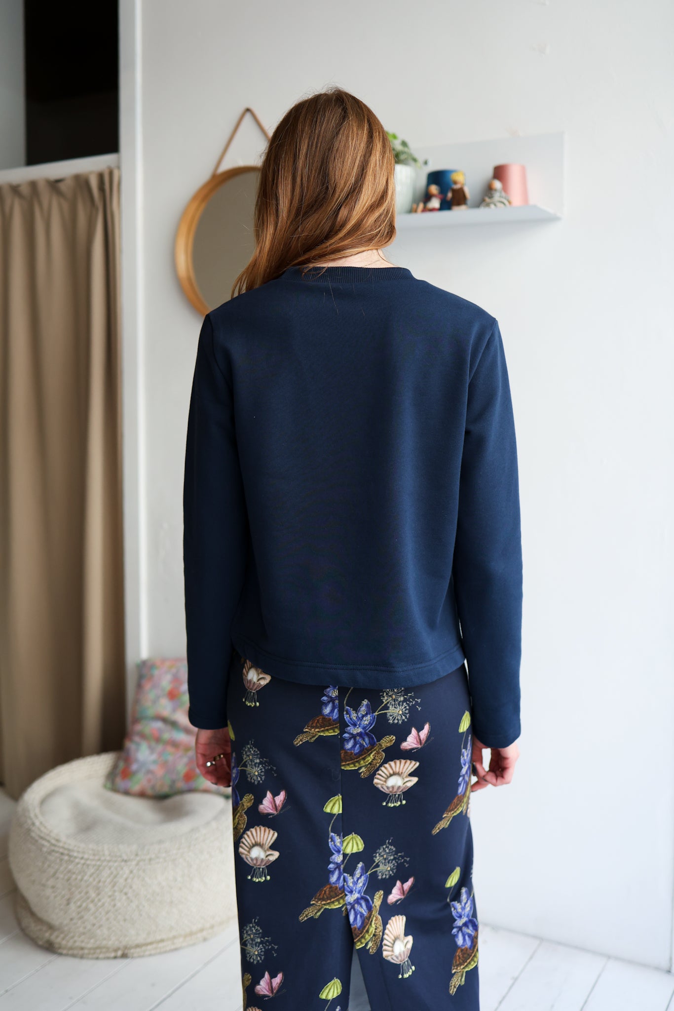 Person wearing a navy sweatshirt and floral pants in a room with a mirror and shelf.