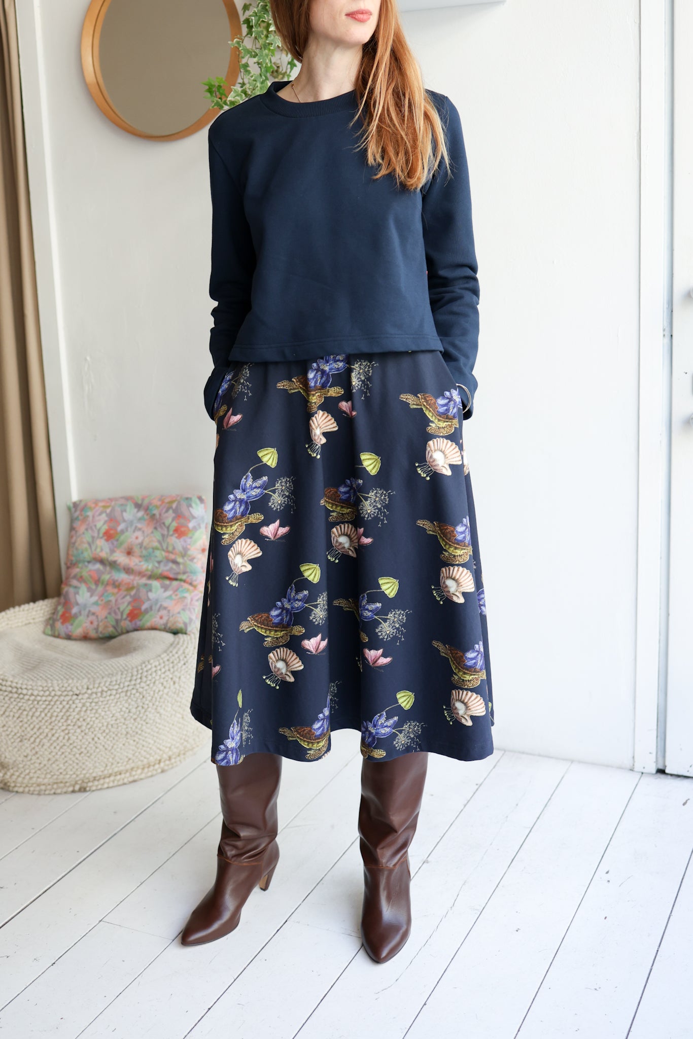 Woman wearing a navy sweater and floral skirt with brown knee-high boots indoors.