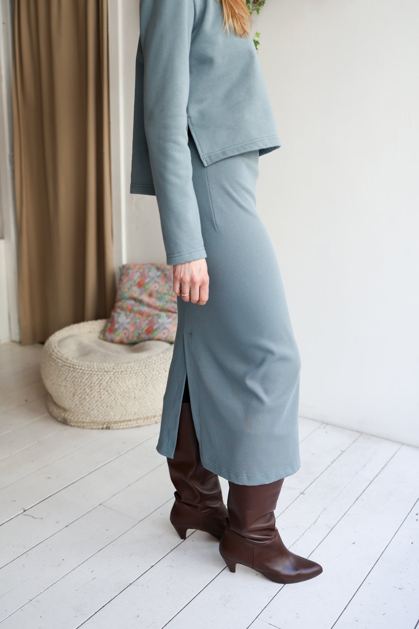 Person wearing a light blue outfit with a skirt and brown boots in a room with a white wall and wooden floor.