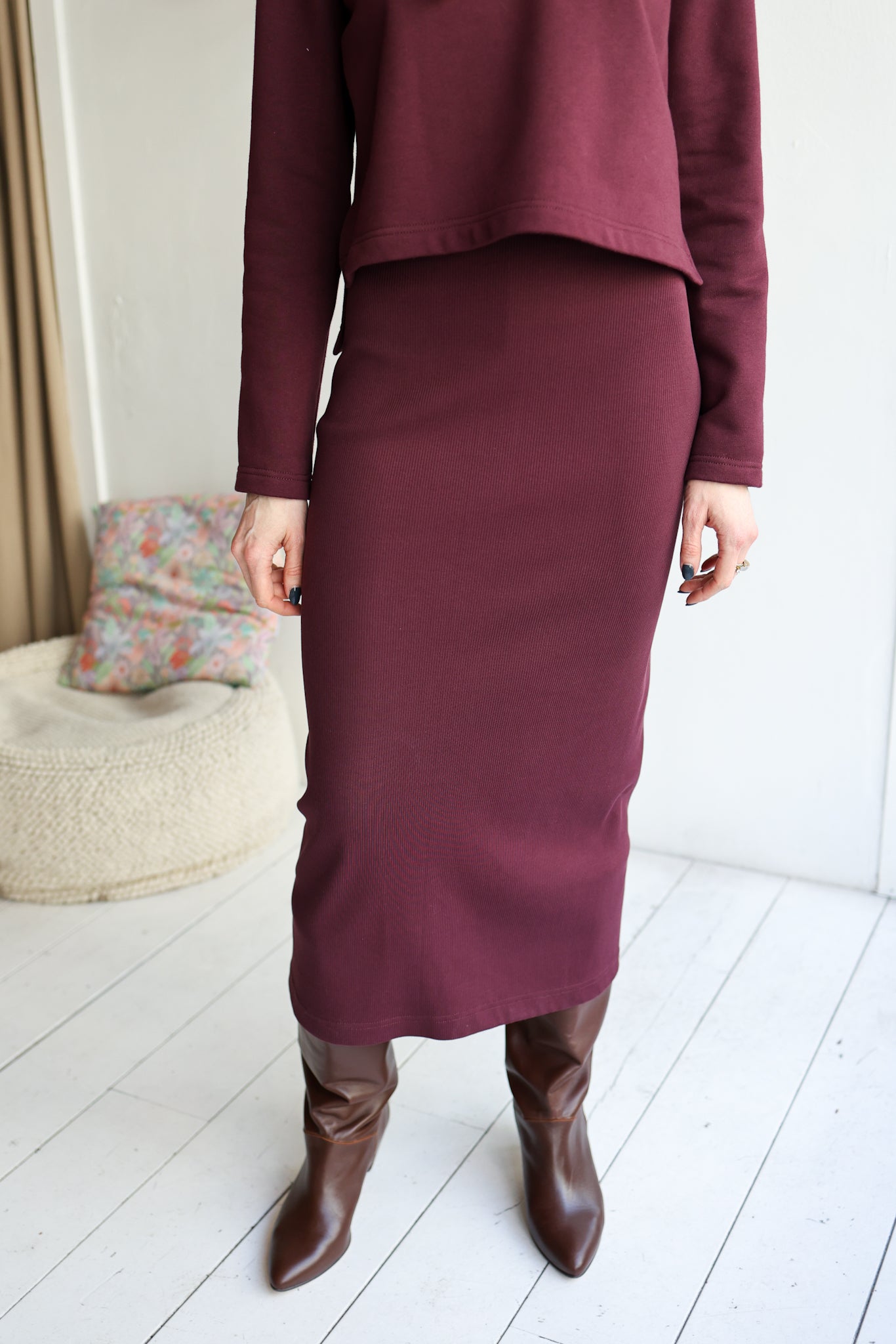 Person wearing a burgundy outfit with a skirt and brown boots in a minimalistic room.
