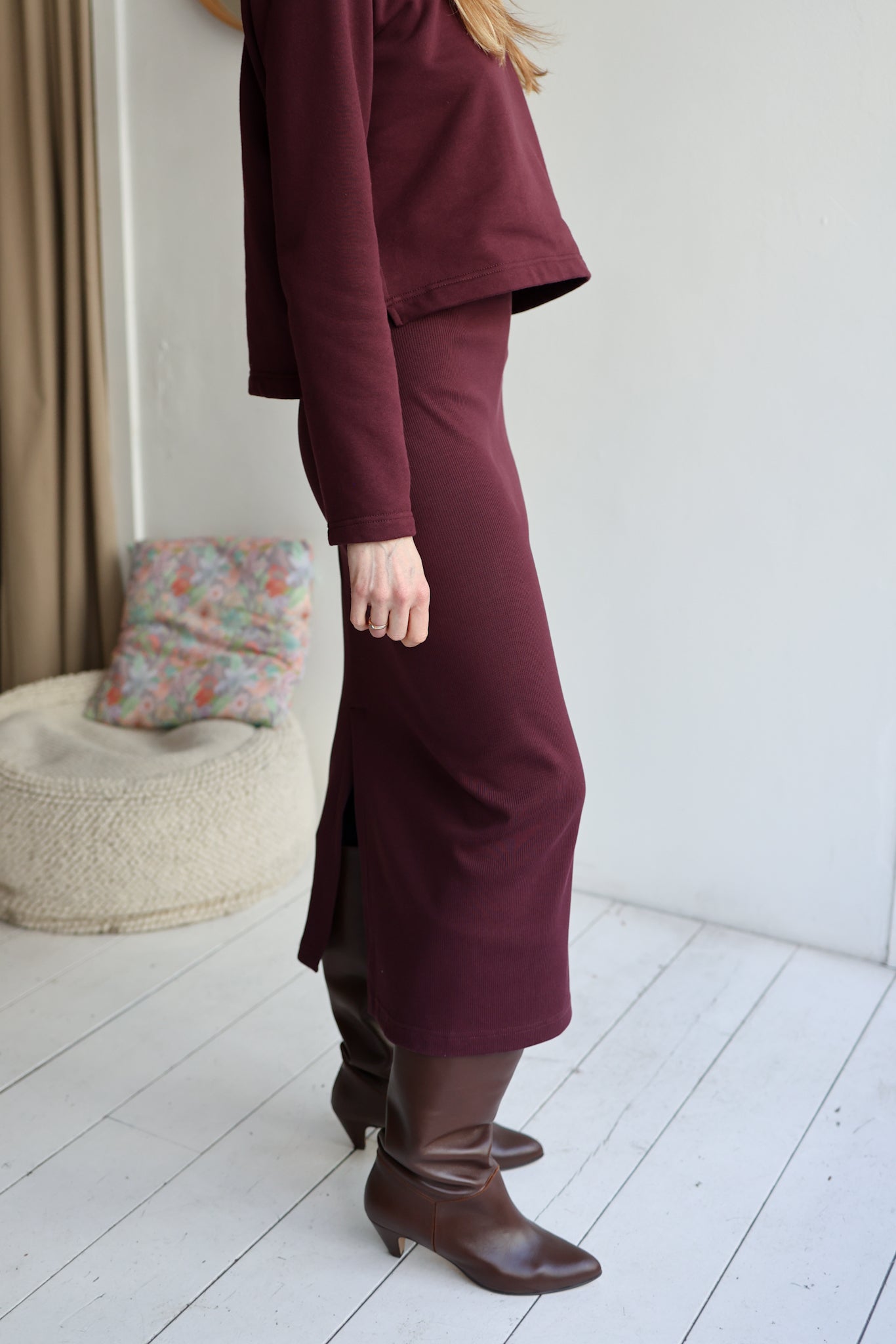Person wearing a burgundy outfit with a skirt and boots in a room with a white wall and wooden floor.