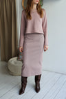 Woman wearing a pink top and skirt with brown boots in a minimalistic room.