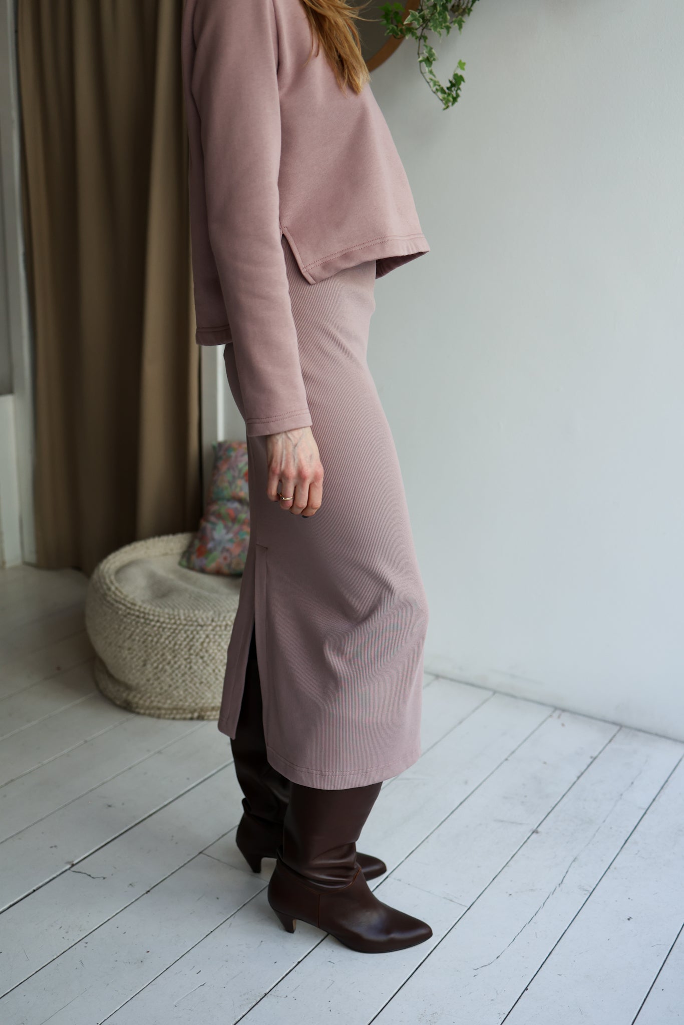 Person wearing a long mauve coat and brown boots in a minimalistic room.