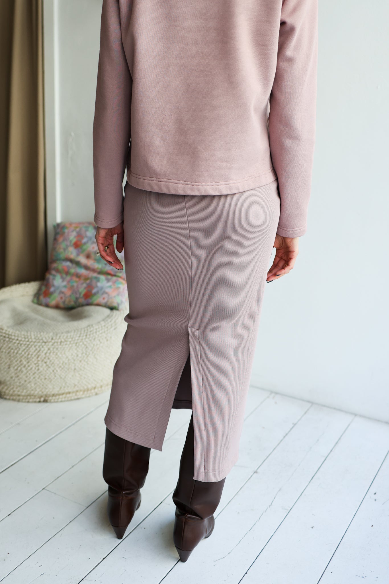 Person wearing a pink sweater and skirt with brown shoes in a minimalistic room.