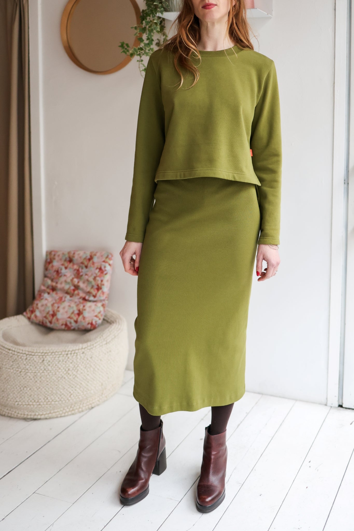 Person wearing a green outfit standing in a room with a white wall and wooden floor.