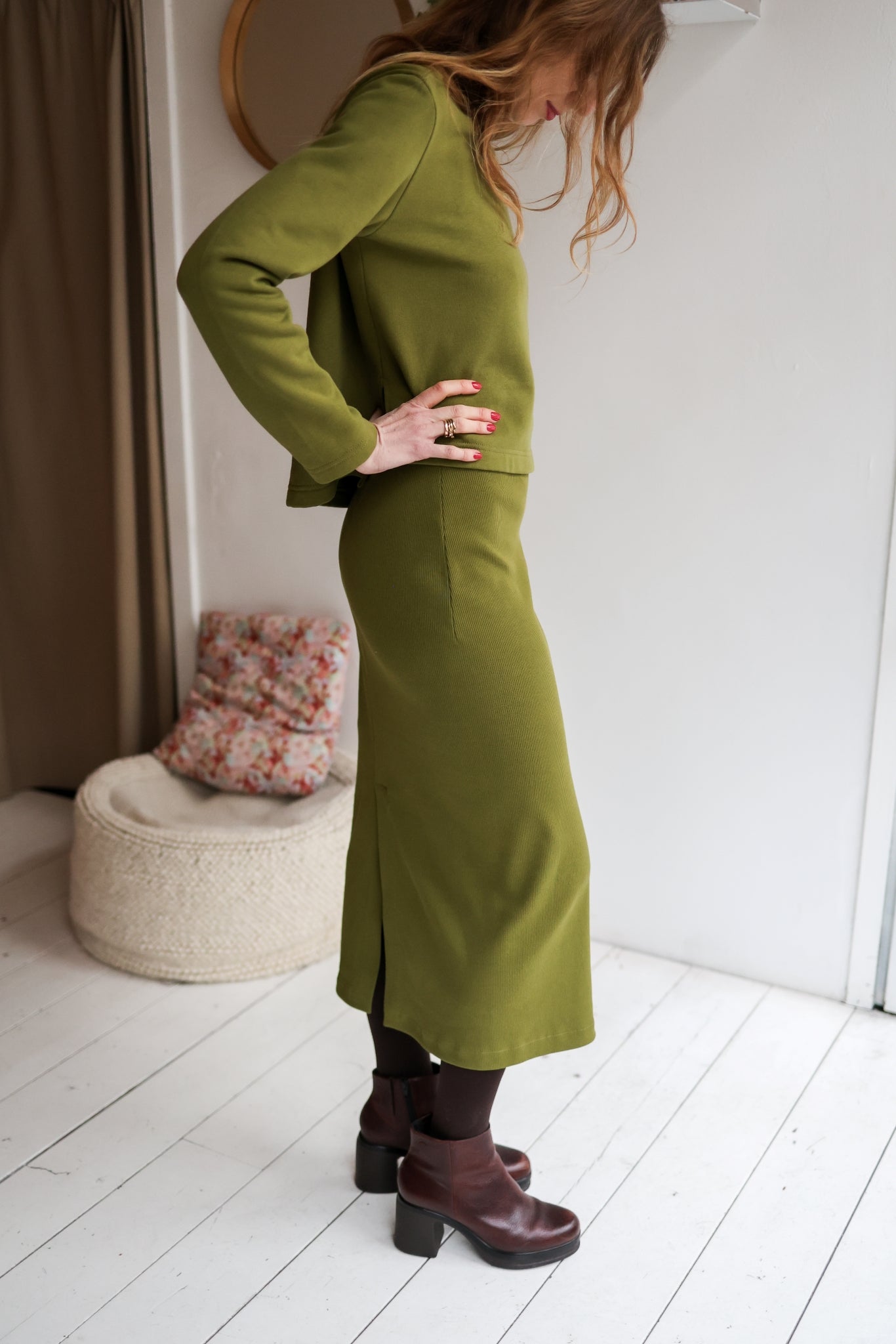 Woman wearing a green outfit standing in a room with a white wall and wooden floor.