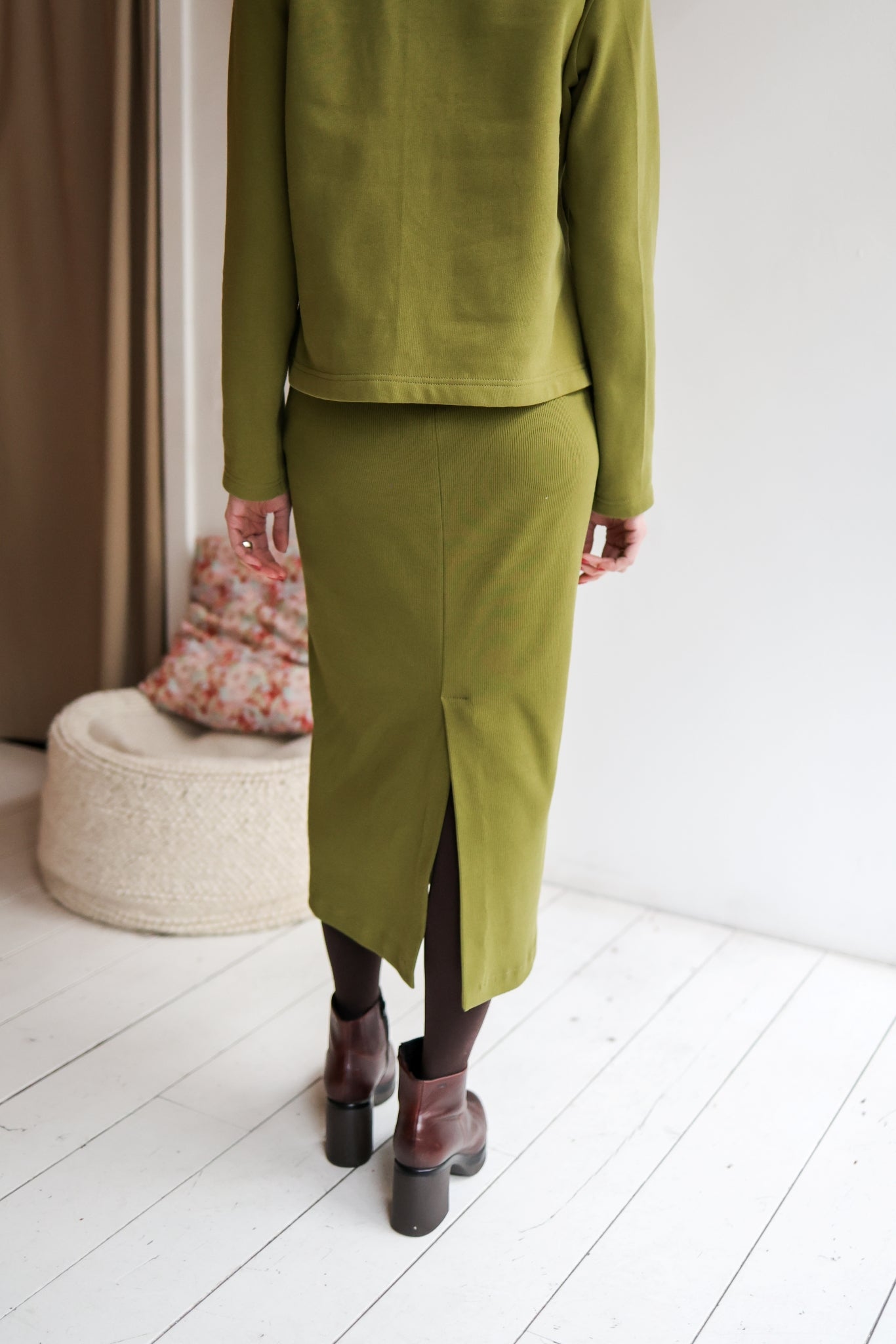 Person wearing a green outfit standing in a minimalistic room with white floor and light-colored walls.