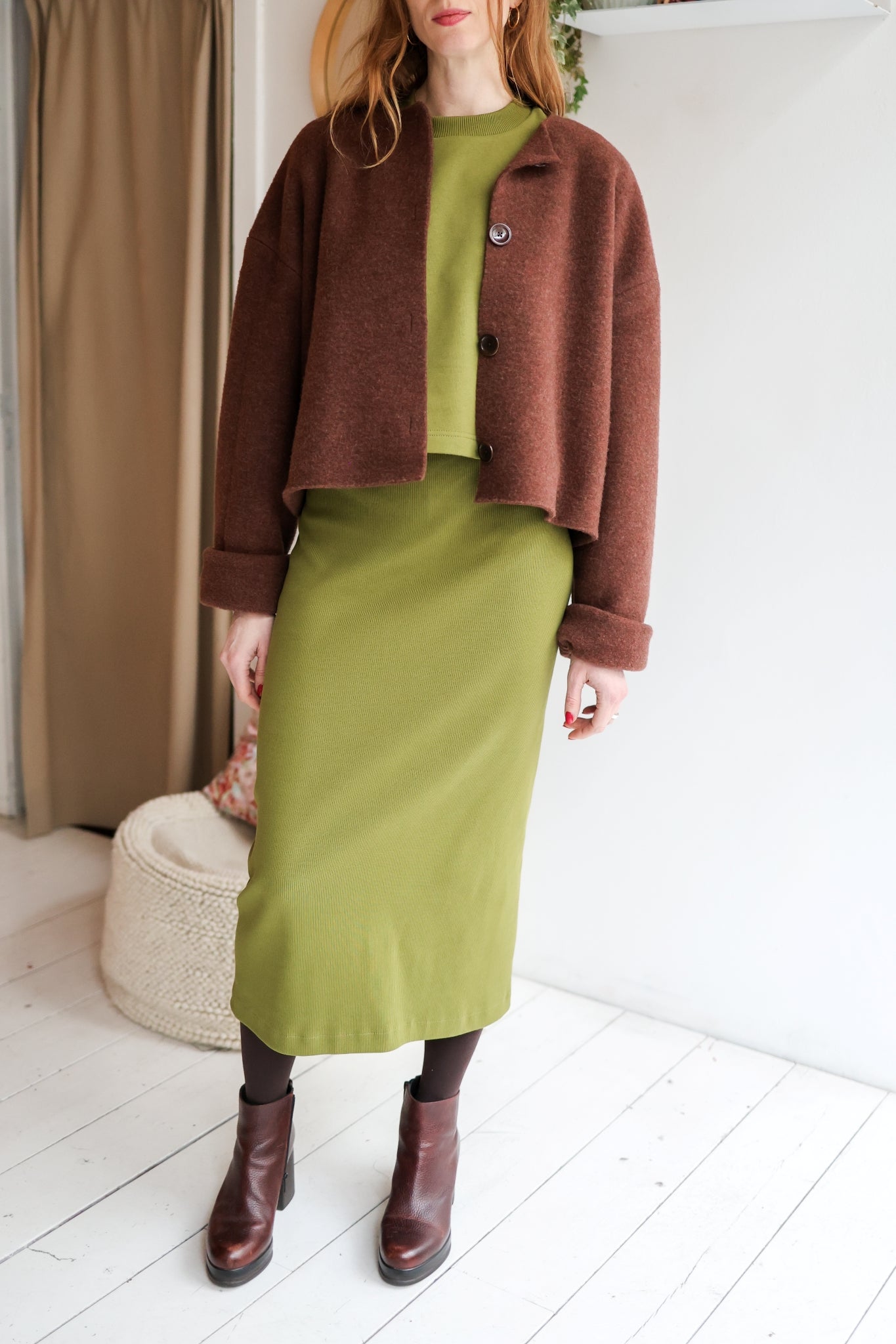 Woman wearing a brown jacket, green top, and green skirt standing in a room with white walls and a beige curtain.