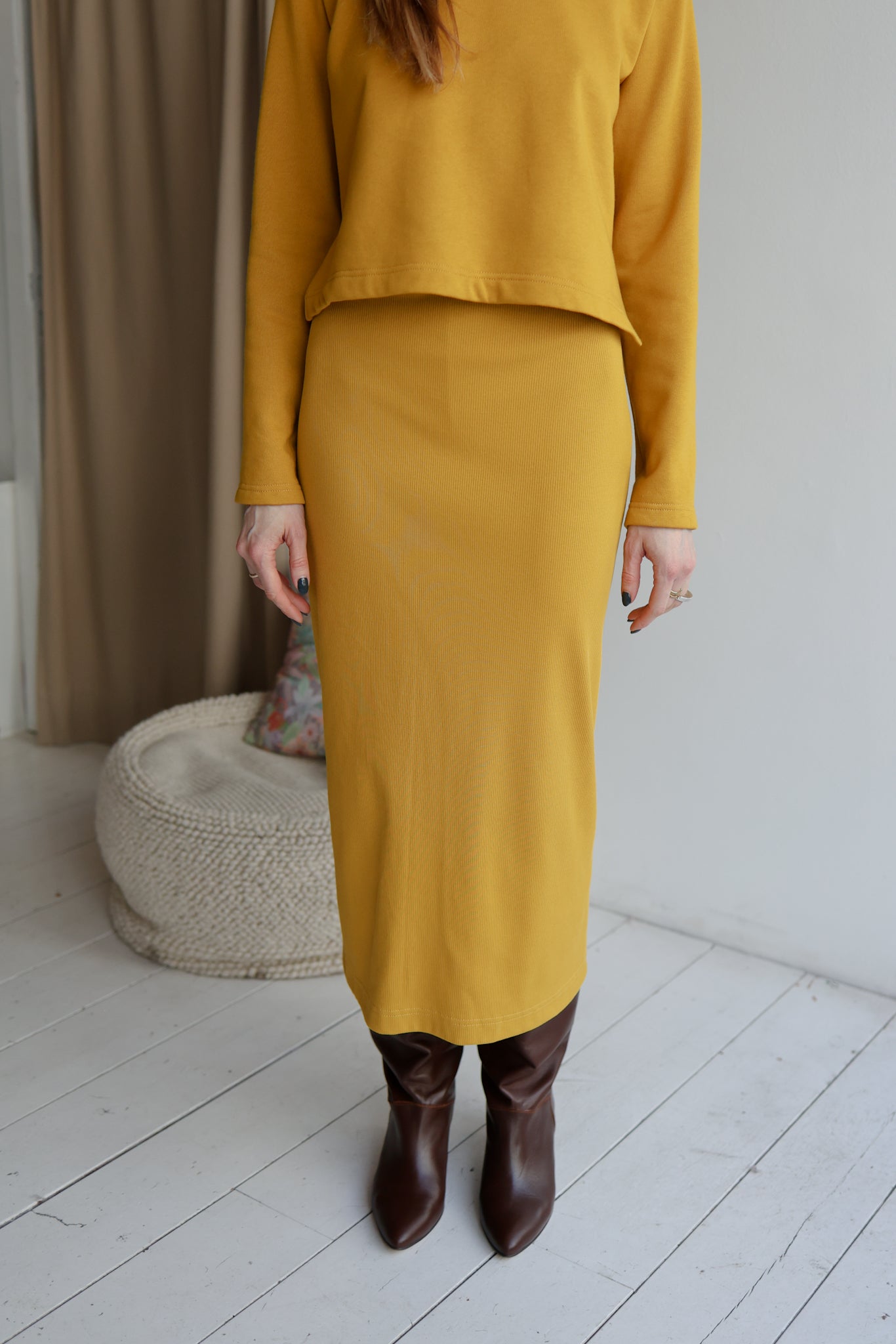 Person wearing a mustard yellow outfit with a skirt and top, standing in a minimalistic room.