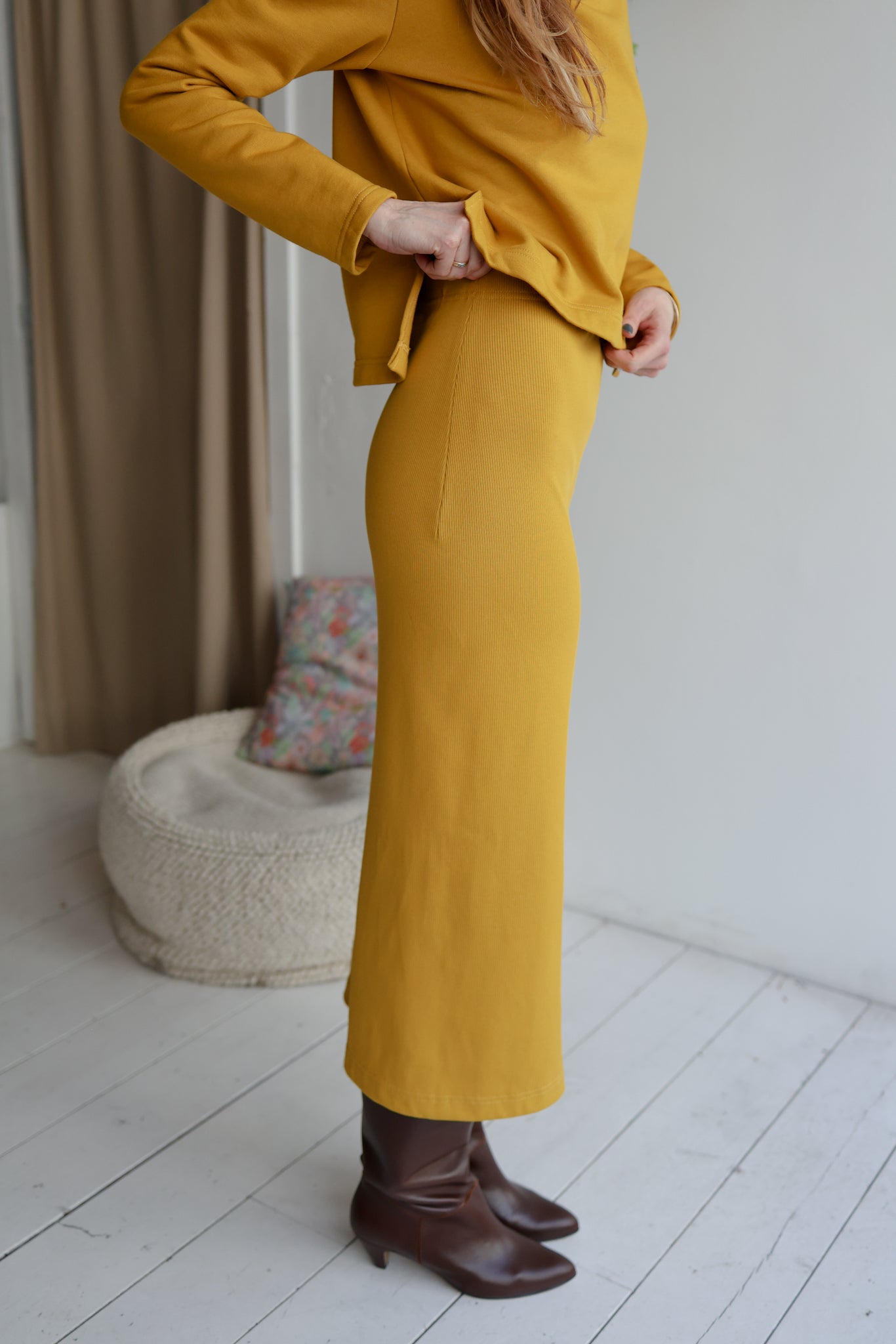 Person wearing a mustard yellow outfit with brown boots in a minimal indoor setting.