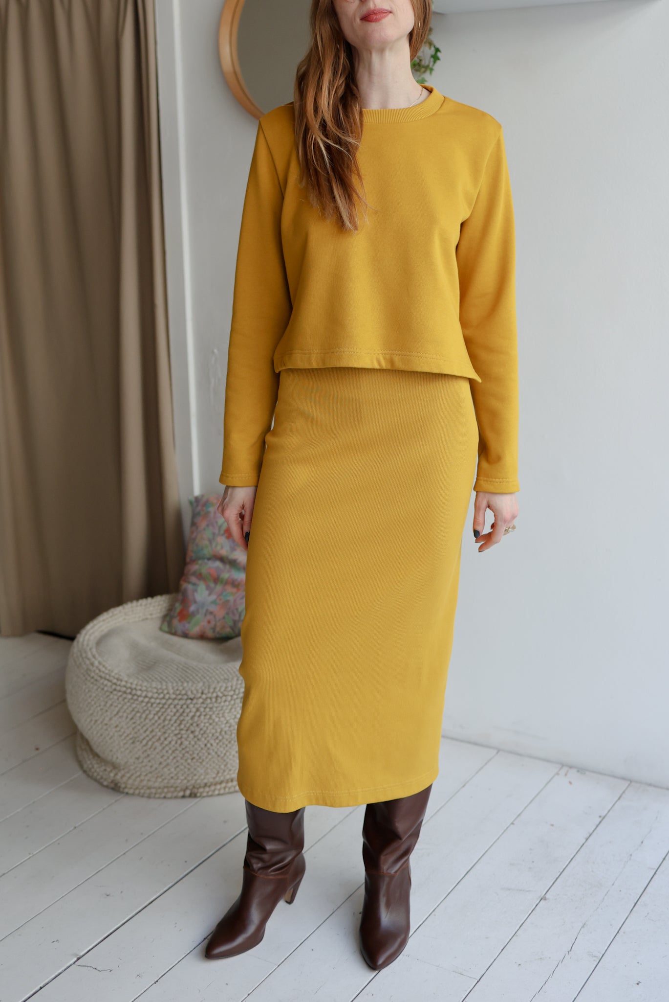 Woman wearing a mustard yellow outfit with a skirt and top, standing in a room with a white wall and brown curtain.
