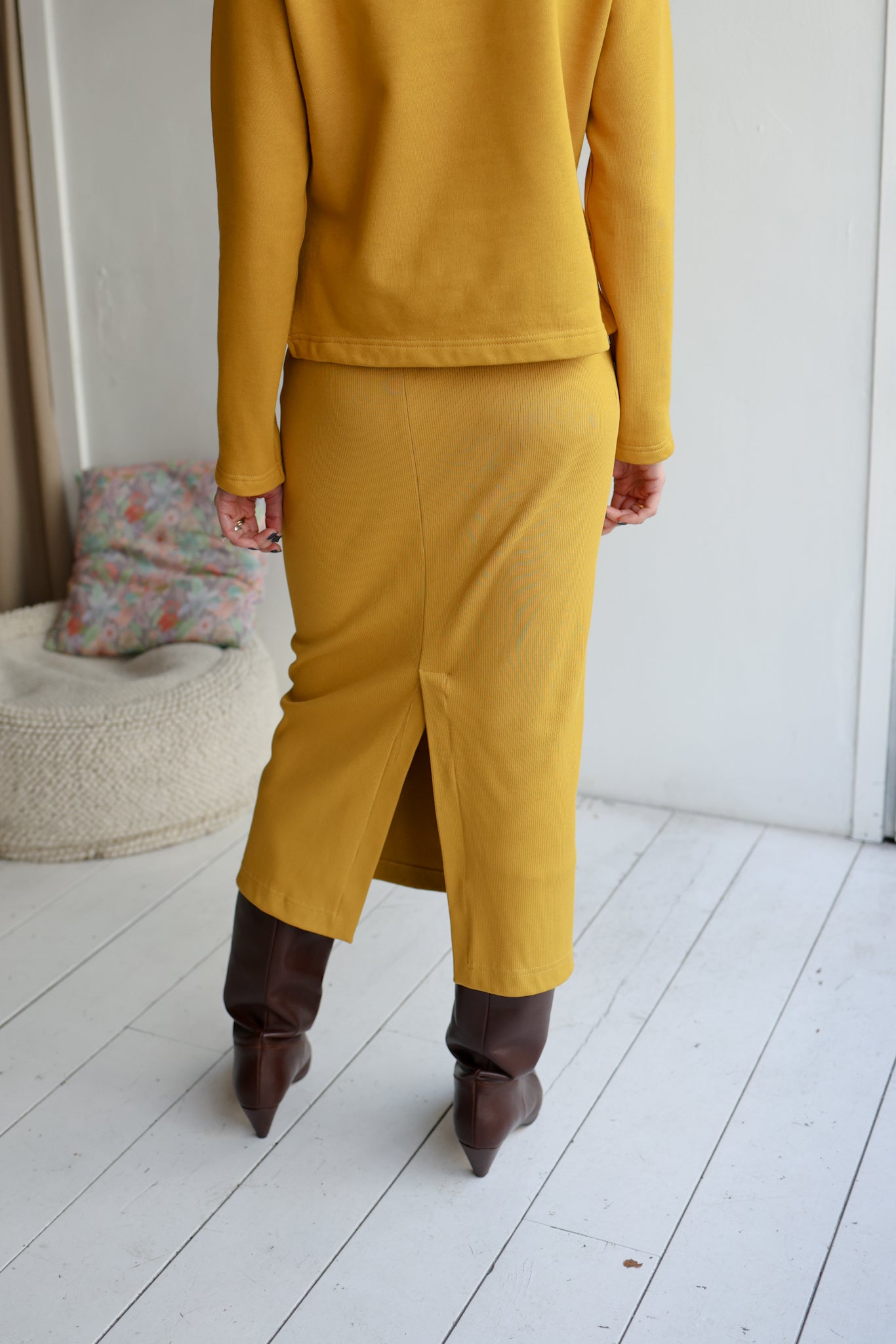 Person wearing a mustard yellow outfit with brown boots in a room with white walls and a textured rug.