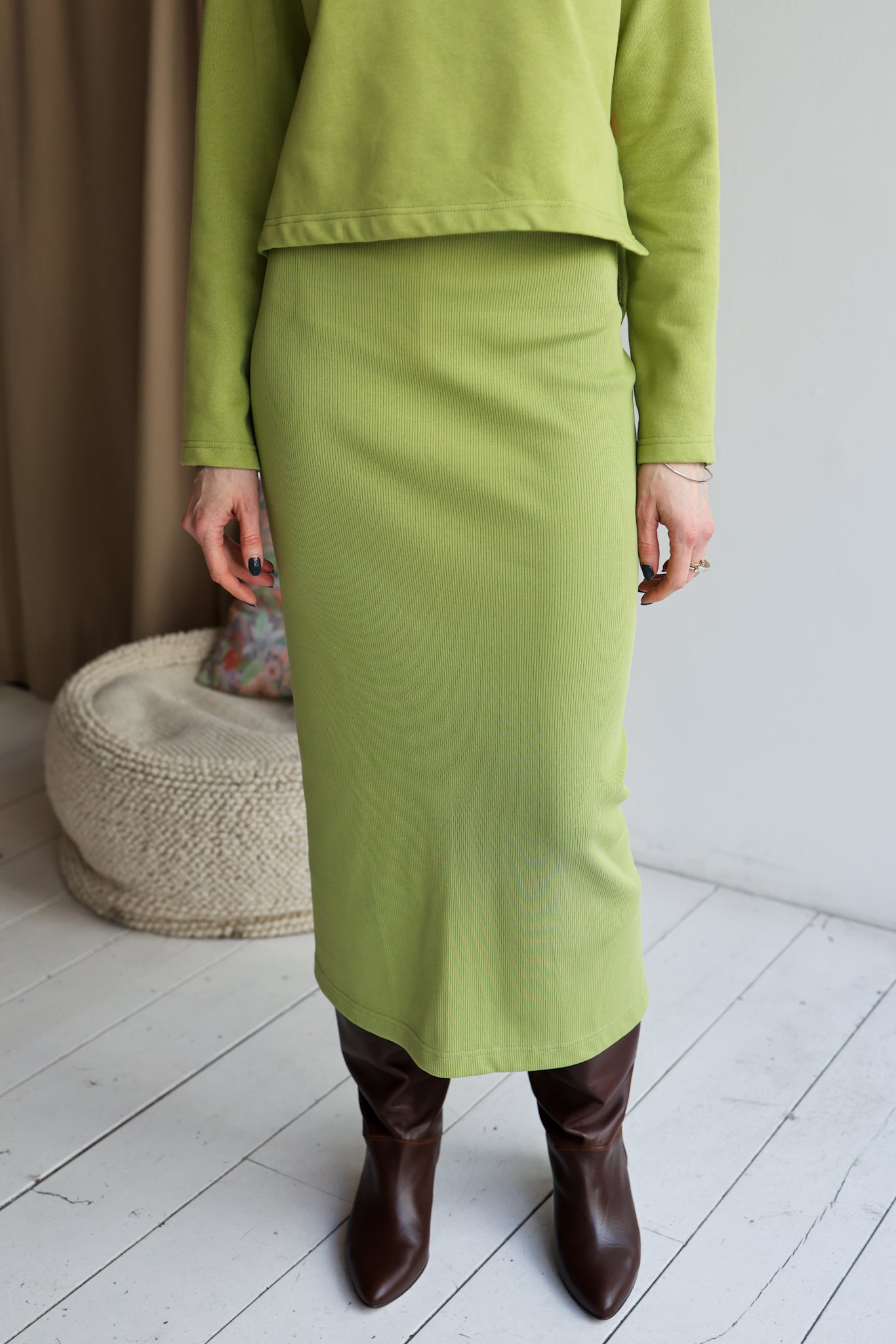 Person wearing a green outfit with a skirt and brown boots in a minimalistic room.