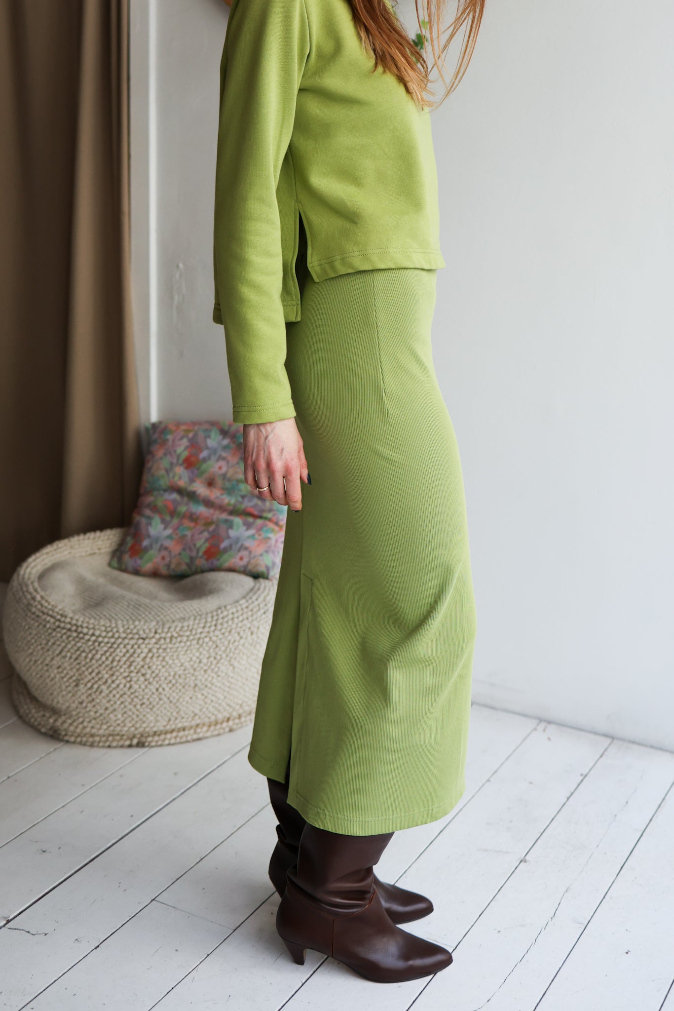 Person wearing a green outfit standing in a room with a white wall and wooden floor.