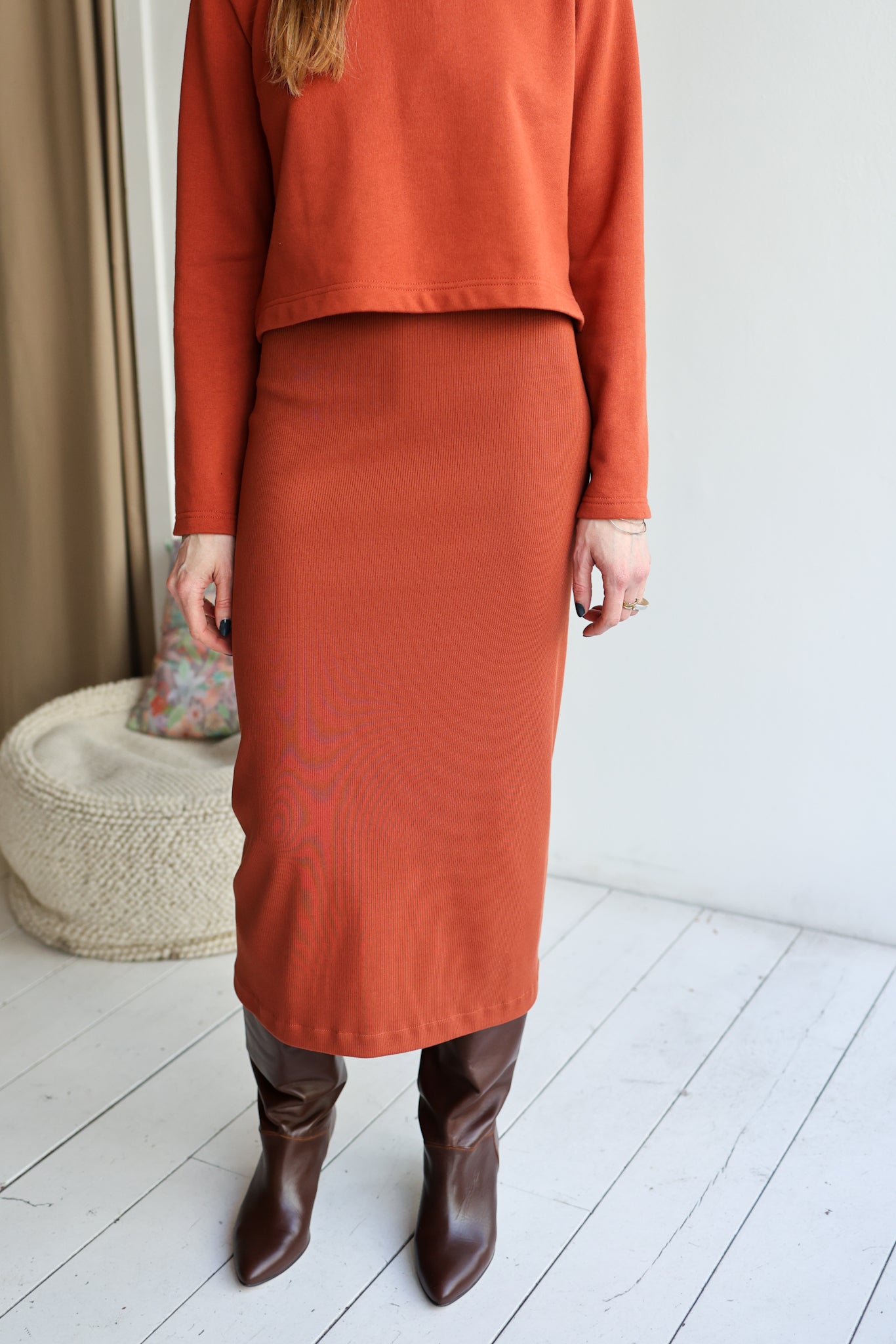 Person wearing a rust-colored outfit with a skirt and boots in a minimal indoor setting.